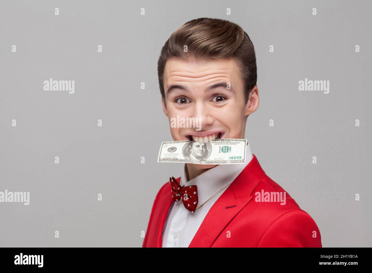 Portrait d'un jeune homme d'affaires cupide avec une coiffure élégante en smoking rouge et noeud papillon tenant un billet de dollar avec des dents, grinçant, concept gourmand en argent. Studio d'intérieur tourné isolé sur fond gris Banque D'Images