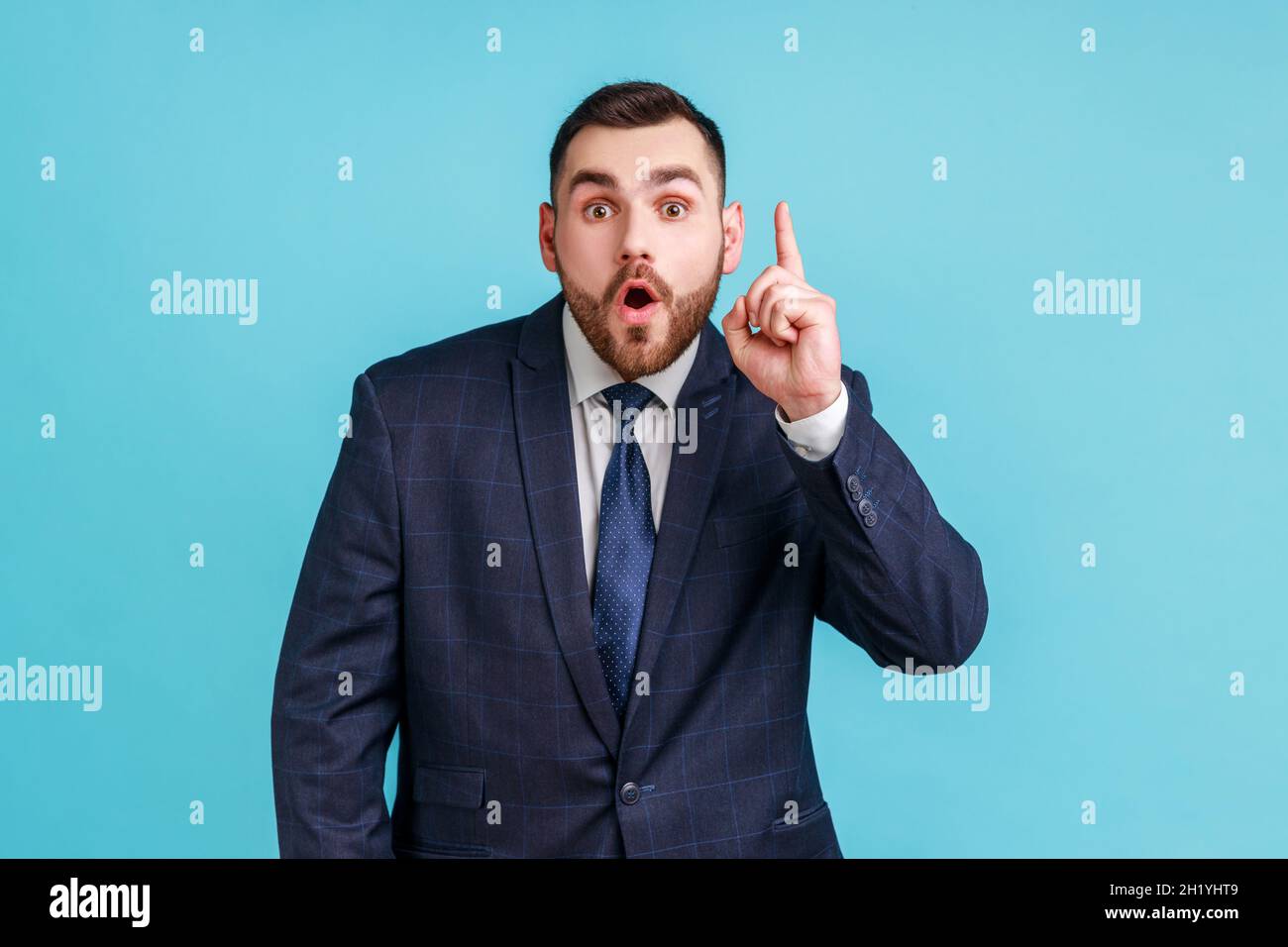 Homme souriant intelligent avec barbe portant un costume de style officiel pointant vers le haut, a une bonne idée de plan de génie, d'inspiration et de créativité.Studio d'intérieur isolé sur fond bleu. Banque D'Images