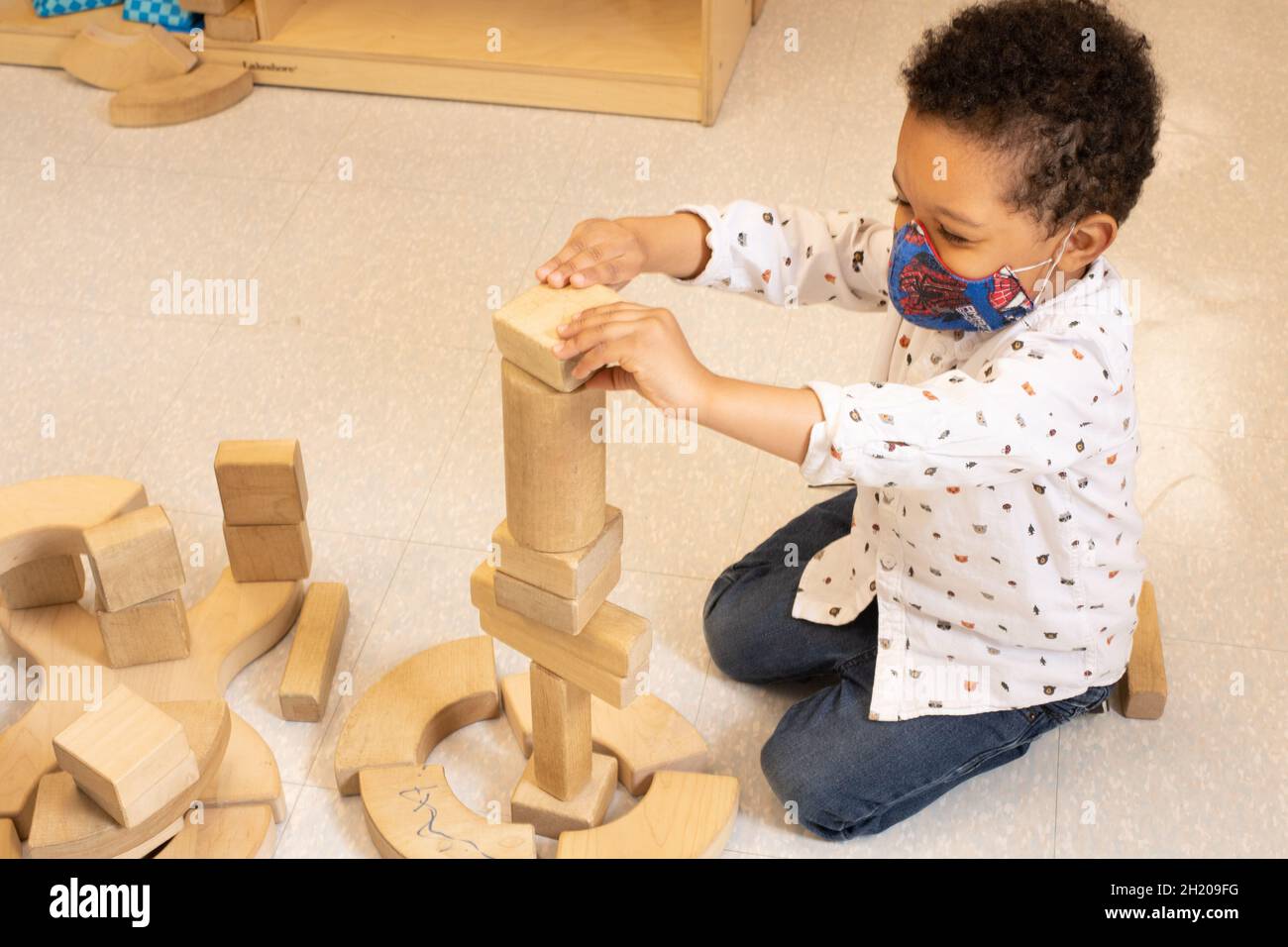 Éducation préscolaire garçon de 3-4 ans jouant dans la zone de bloc mettant bloc en bois sur le dessus de la tour qu'il a construit, portant un masque facial pour protéger de Covid-19 Banque D'Images