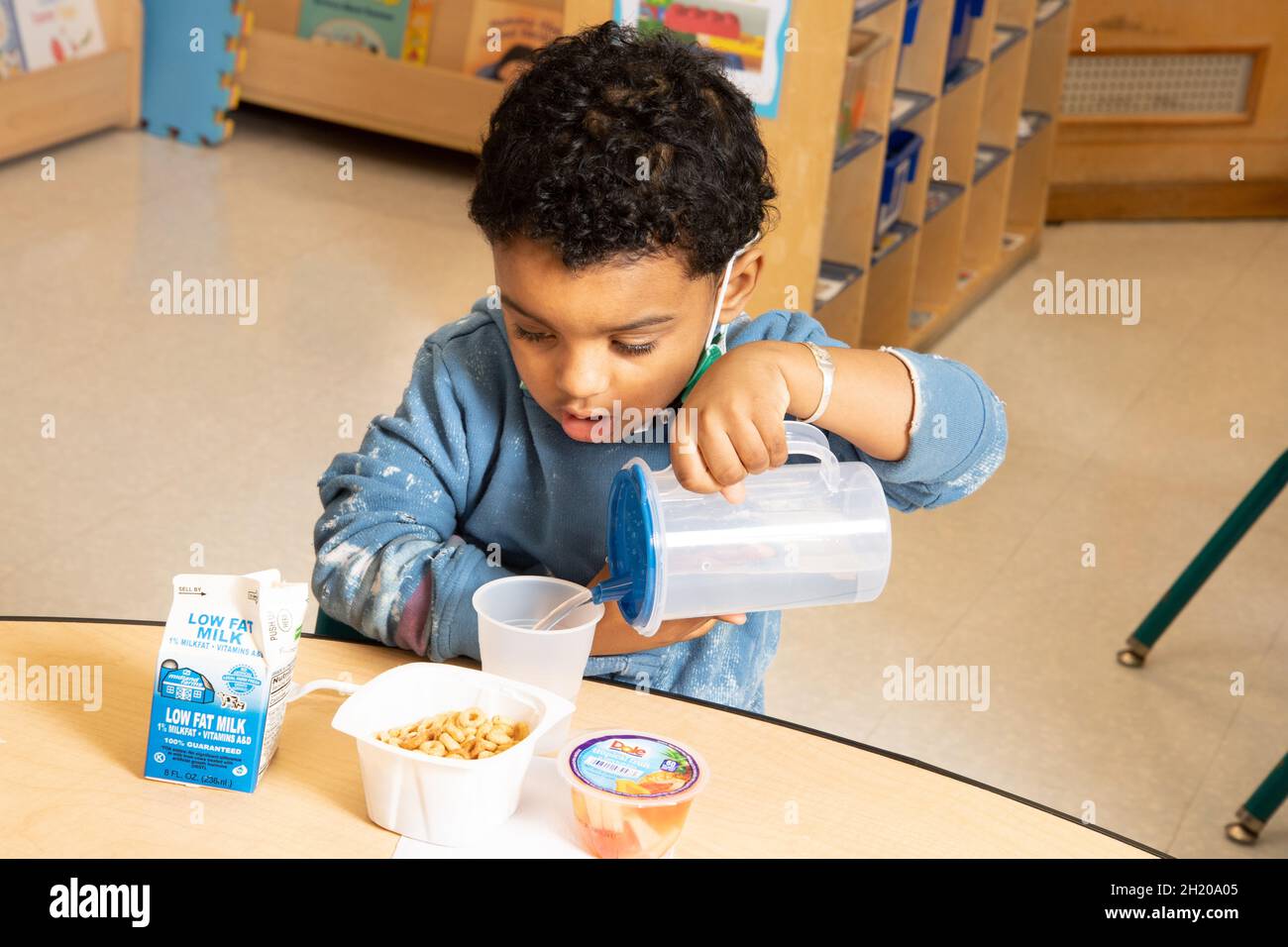 Éducation préscolaire garçon de 3-4 ans versant de l'eau au petit déjeuner, de la cérale, du lait et des fruits sur la table avant lui Banque D'Images
