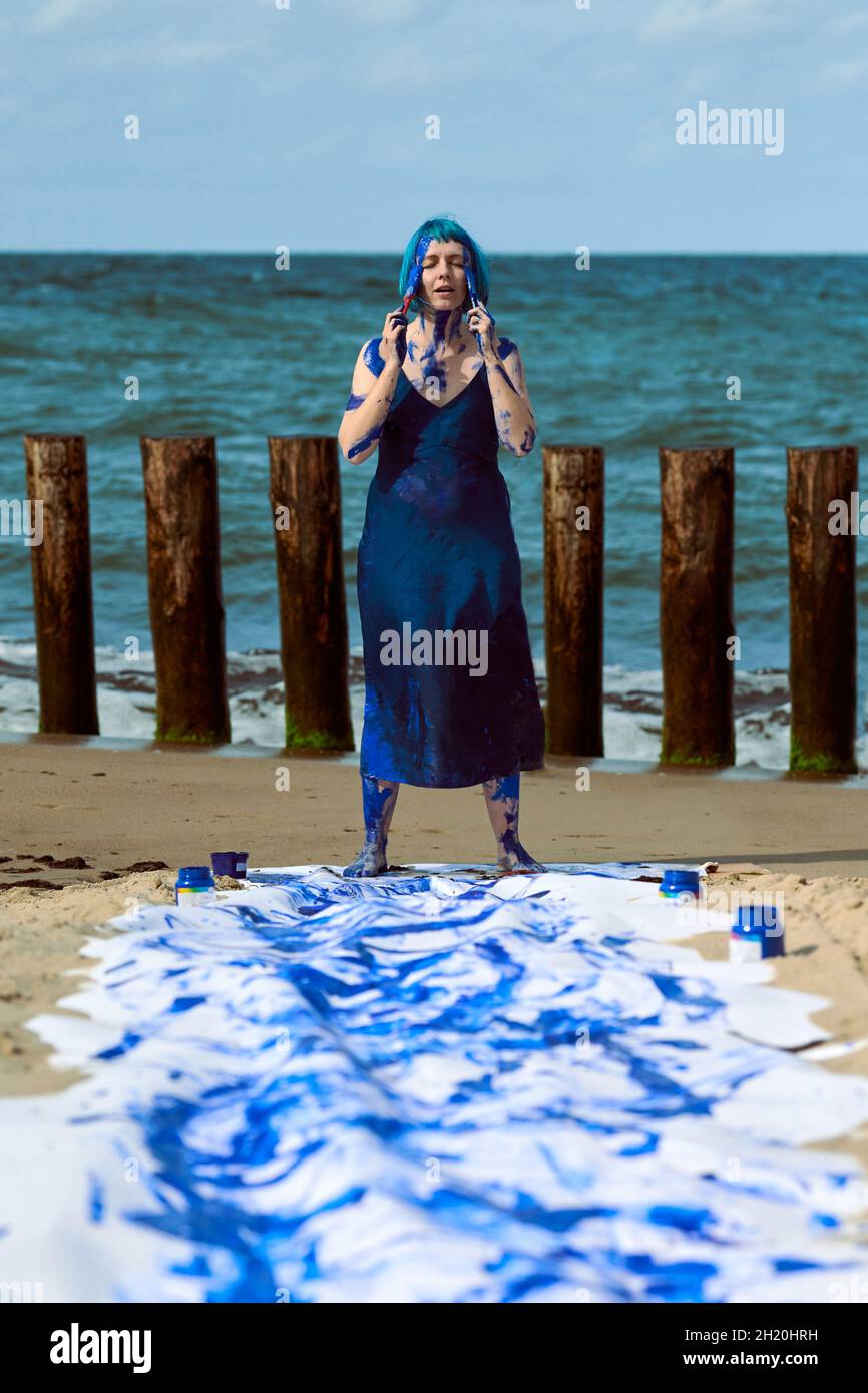 Jeune femme artiste de performance en robe bleu foncé étalé de peintures bleu gouache dansant heureusement les mains sur la plage.Toile pour peinture expressive W Banque D'Images