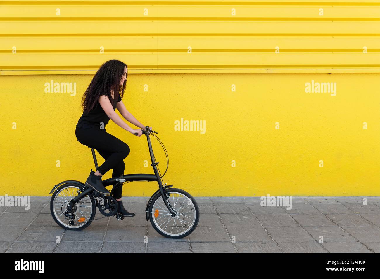 Jeune femme noire aux cheveux longs et tenue noire à cheval sur son vélo devant Un mur jaune Banque D'Images