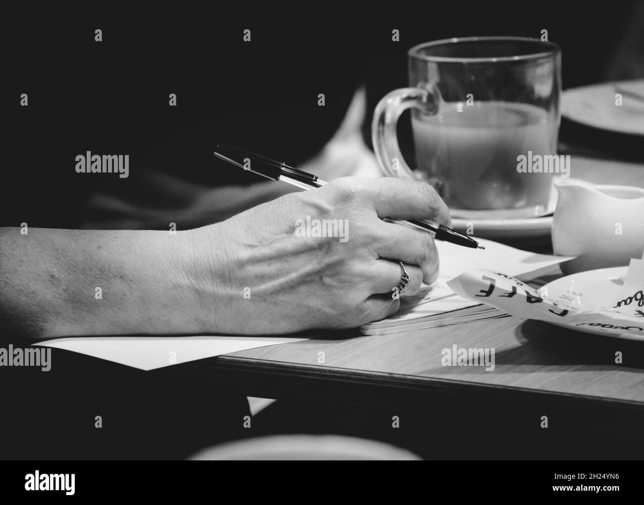 Photographie en noir et blanc d'une femme écrivant à la main pendant la pause, avec café et assiette. Banque D'Images