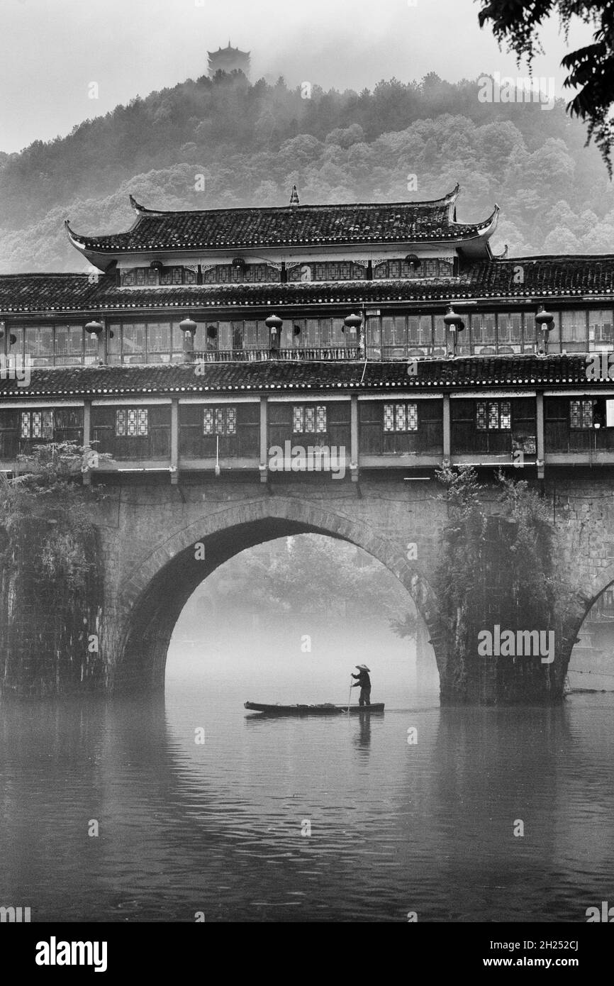 Un homme pagaie un sampan dans le brouillard près du Phoenix Hong Bridge sur la rivière Tuojiang, Fenghuang, Chine. Banque D'Images