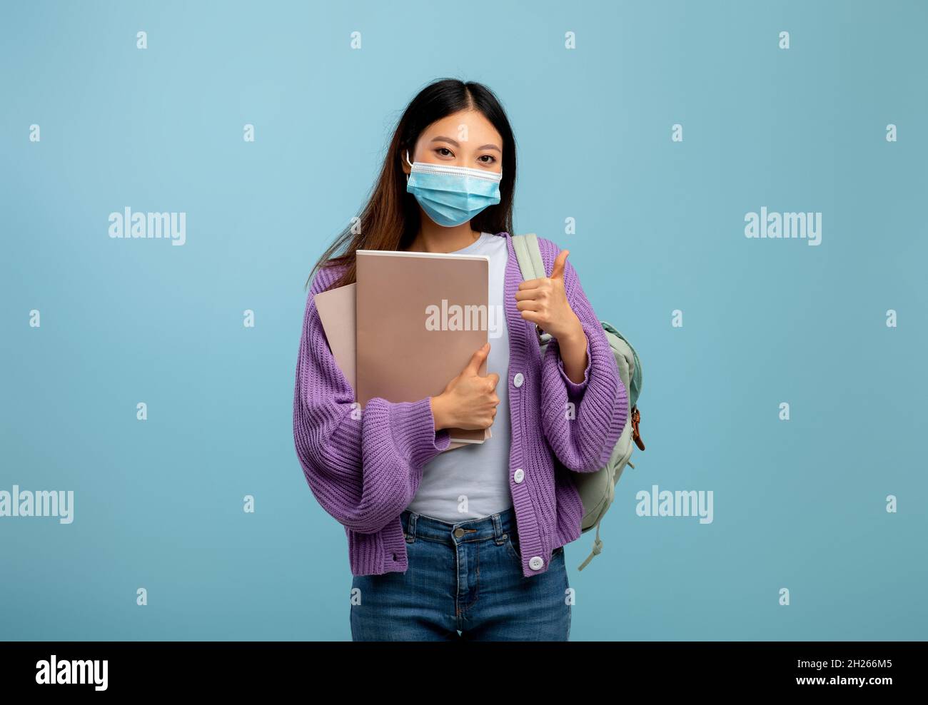 Femme asiatique étudiant en masque avec sac à dos tenant des livres et montrant le pouce vers le haut, debout sur fond bleu Banque D'Images