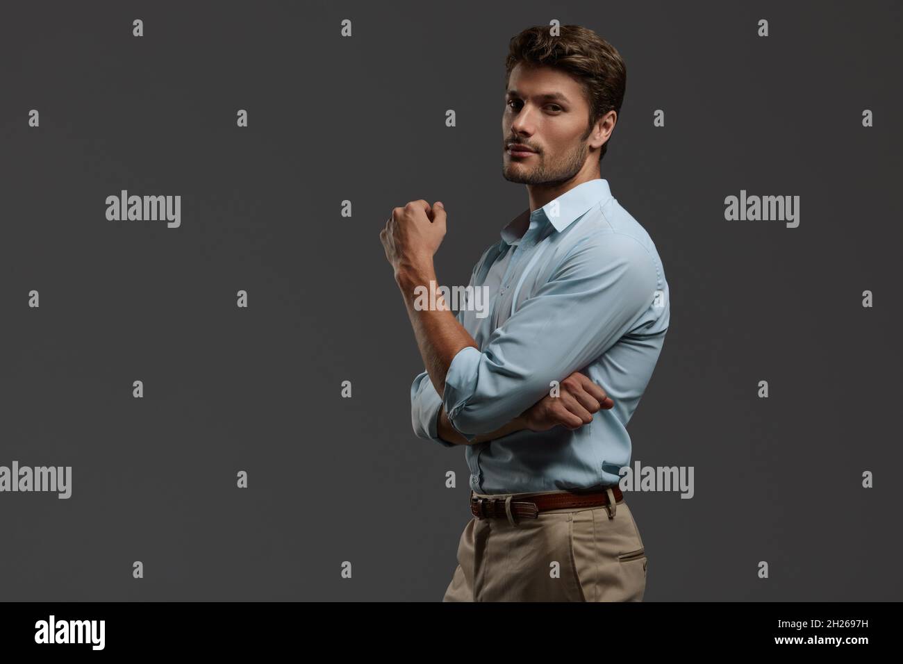 Jeune homme d'affaires européen avec confiance et bras croisés.Vue latérale d'un homme barbu aux cheveux foncés porter une chemise bleue.Isolé sur fond gris foncé Banque D'Images