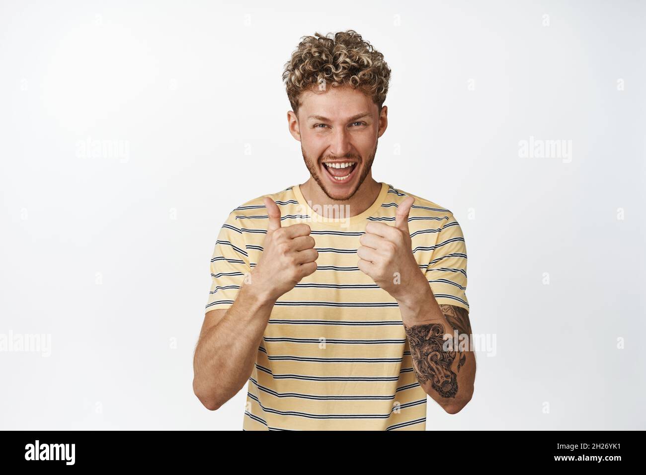 Joyeux homme blond riant et souriant, montrant les pouces vers le haut dans l'approbation, comme quelque chose de bon, en disant oui, debout sur fond blanc Banque D'Images