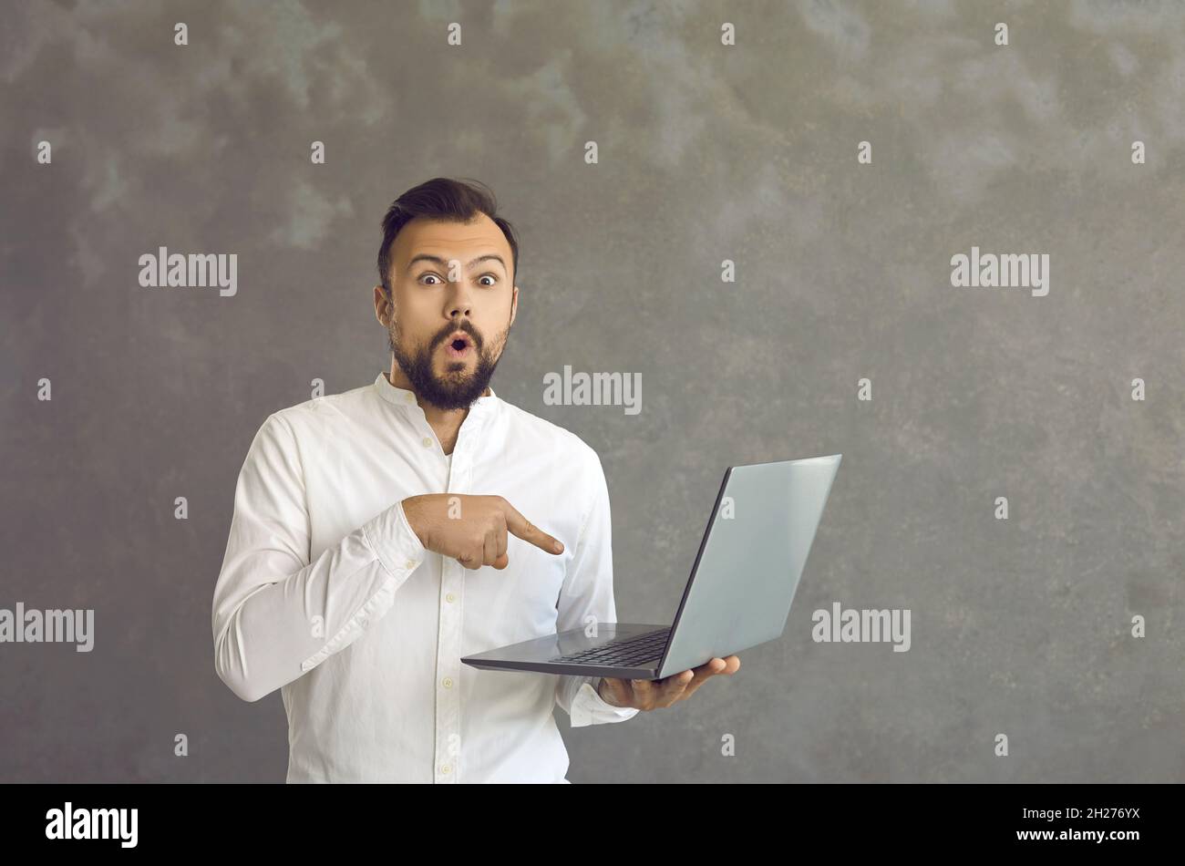 Un homme choqué pointe un doigt sur son ordinateur portable, qu'il tient dans sa main debout sur un fond gris. Banque D'Images