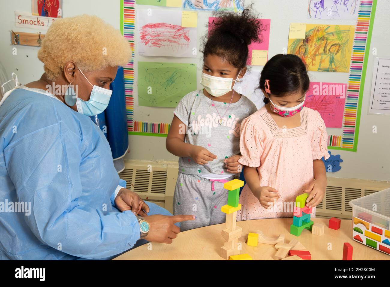 Éducation préscolaire 4-5 ans deux filles bâtiment avec des blocs de bois colorés, enseignant dans la robe de protection leur parler de leur travail Banque D'Images