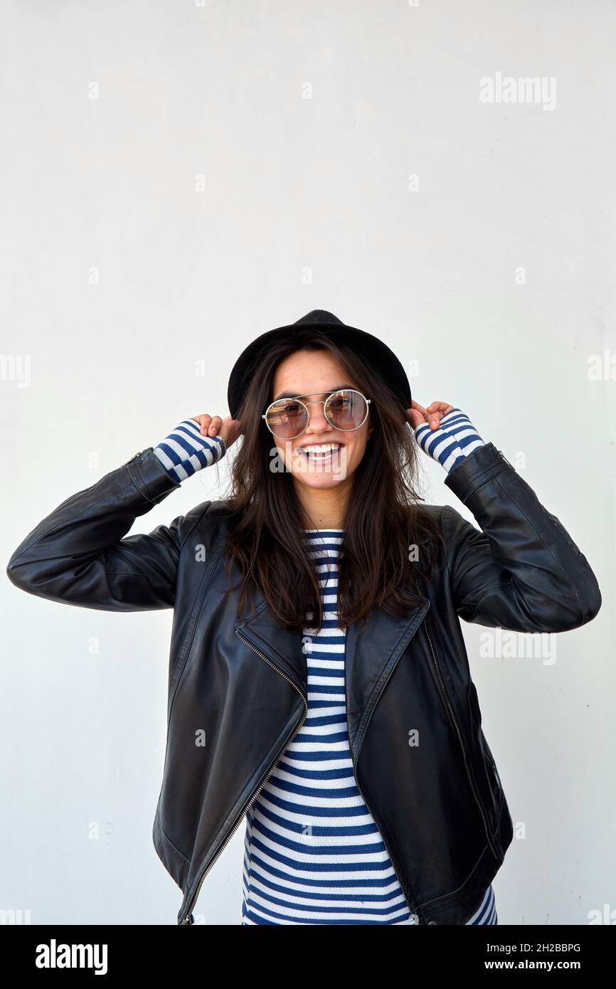 Femme ravie dans une veste en cuir noir et des lunettes de soleil touchant le chapeau et regardant l'appareil photo sur fond blanc Banque D'Images