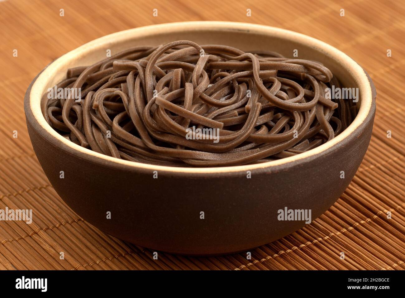 Nouilles soba cuites dans un bol rustique en céramique brune sur un mat de bambou. Banque D'Images