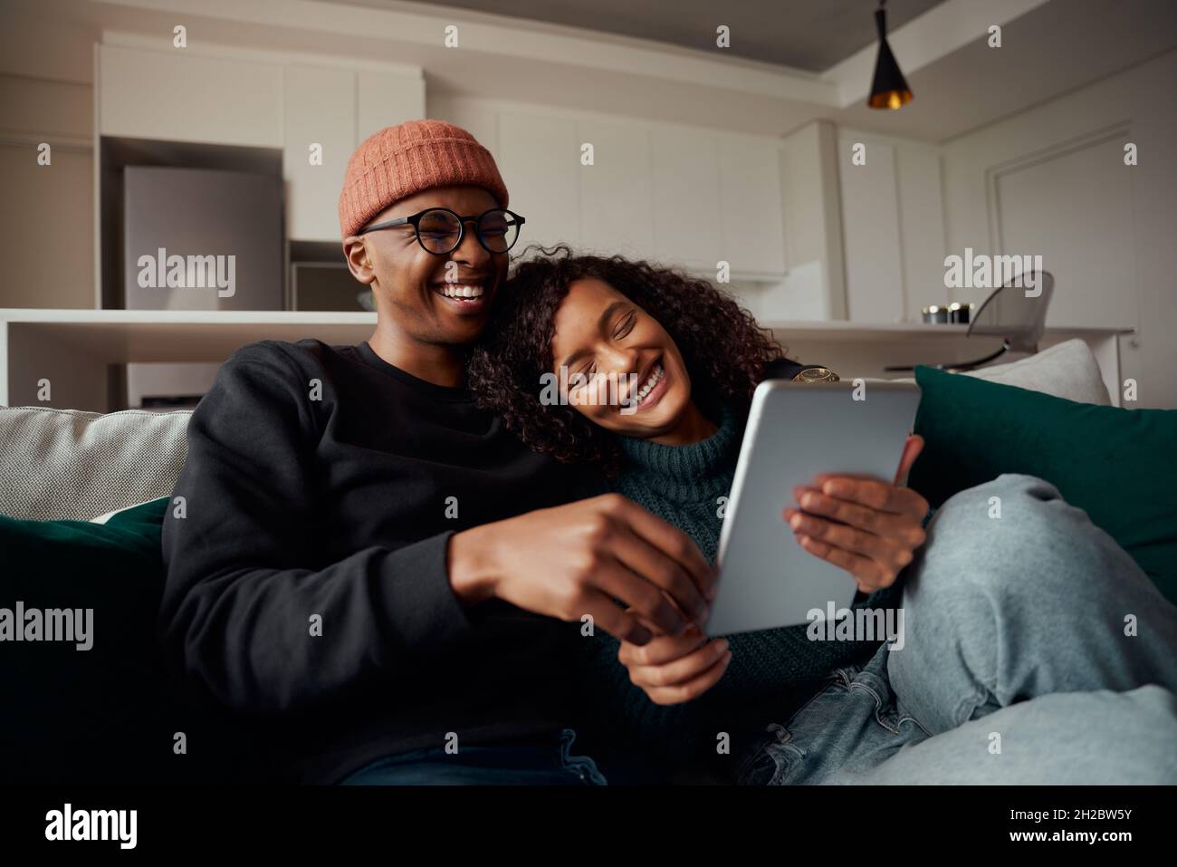 Couple d'adultes branché, multi-ethnique, riant à la vidéo sur tablette sur le canapé dans l'appartement moderne Banque D'Images