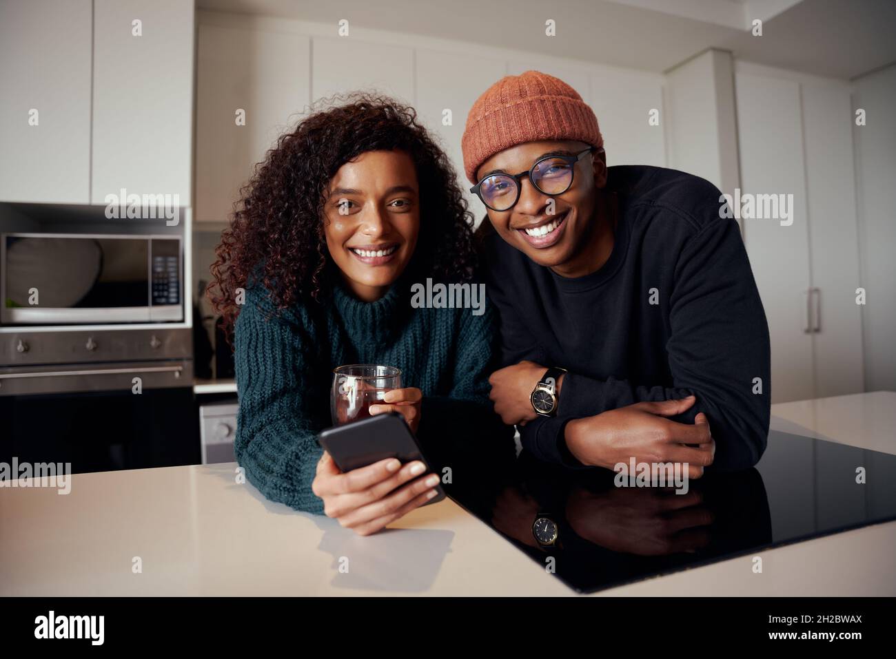 Couple multiethnique souriant à la caméra pendant que dans la cuisine dans l'appartement moderne utilisant le téléphone portable Banque D'Images