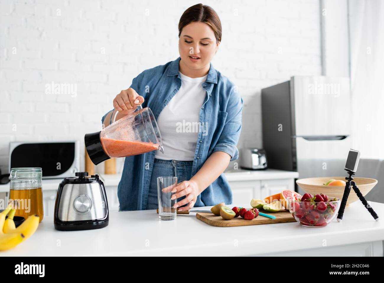 Jeune femme avec un smoothie aux fruits en surpoids près d'un smartphone dans la cuisine Banque D'Images