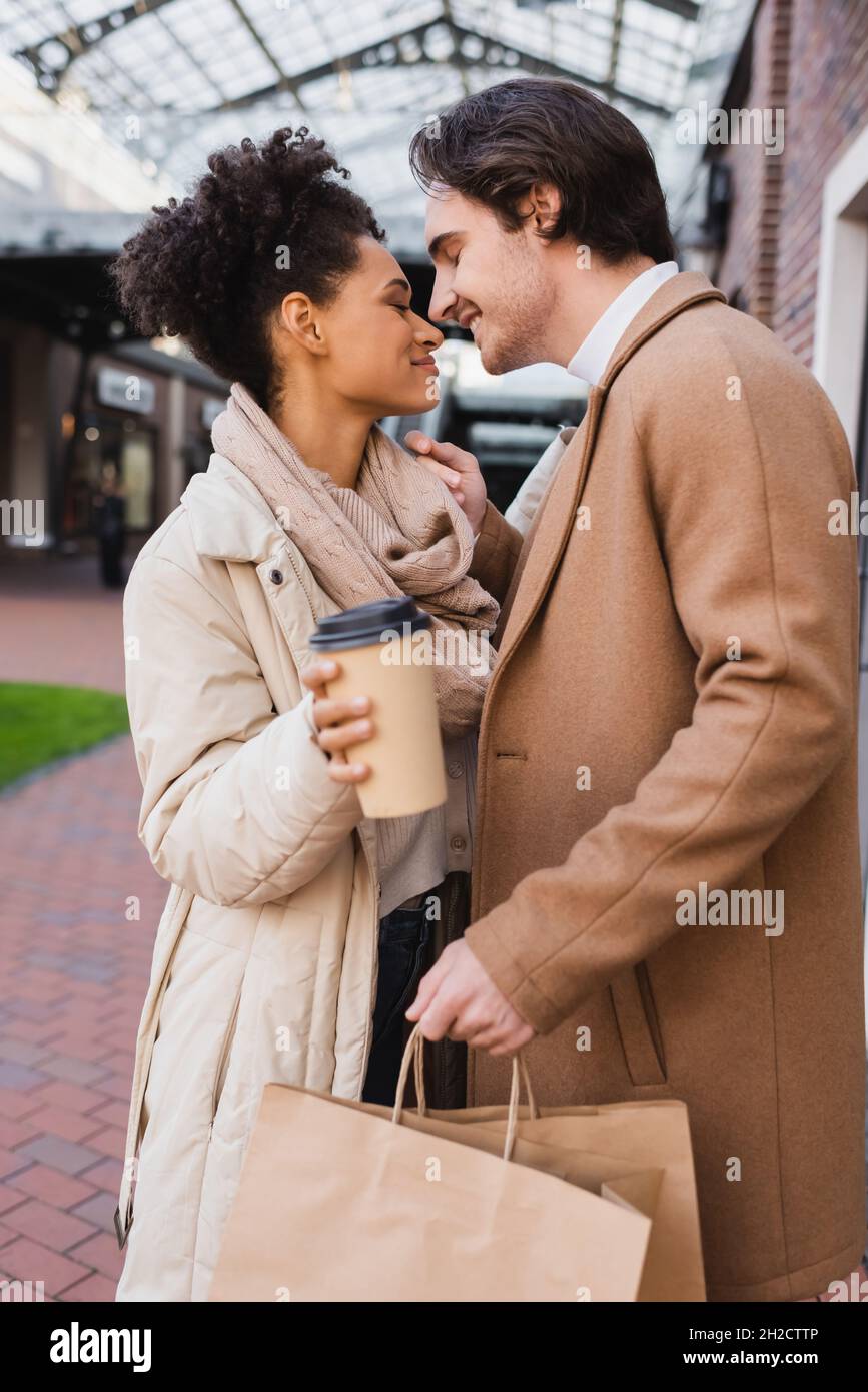 vue latérale d'une femme afro-américaine heureuse avec café à proximité d'un ami heureux tenant des sacs de shopping Banque D'Images