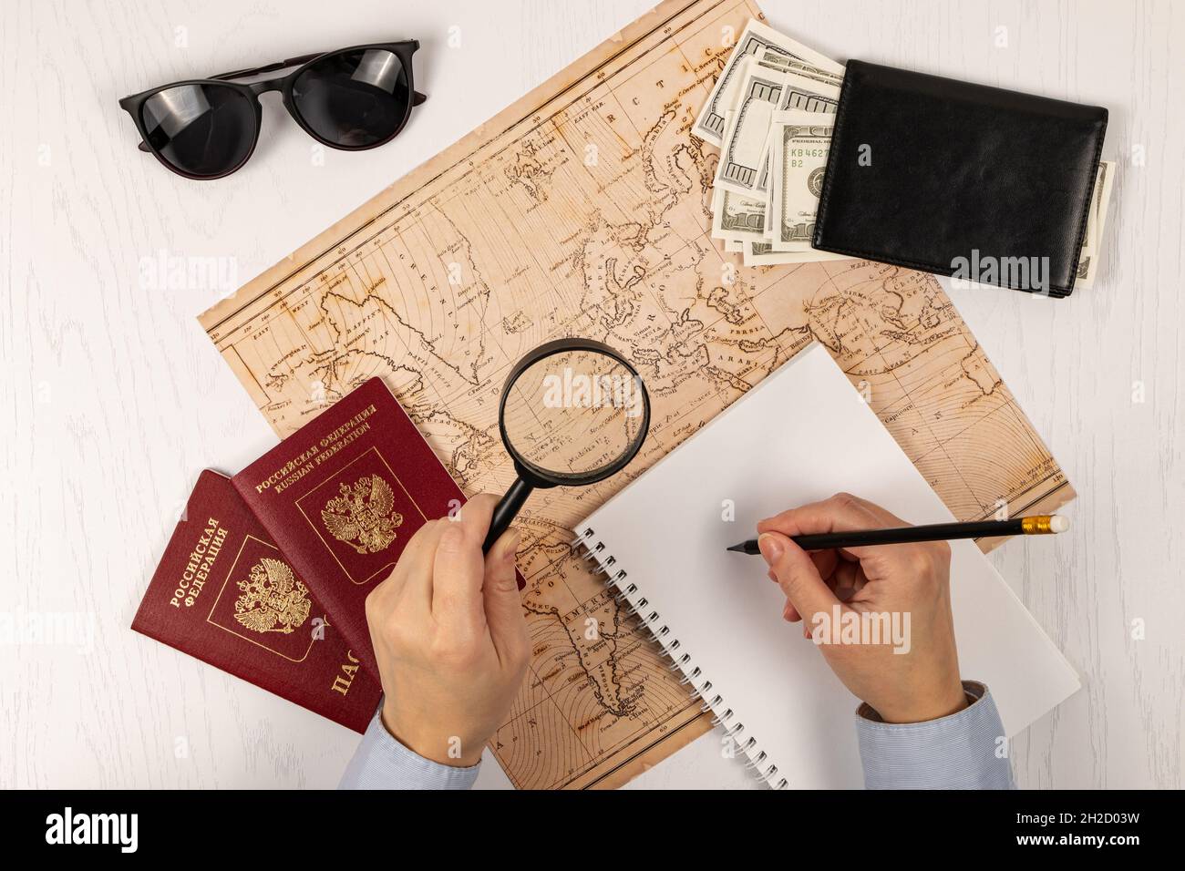la main féminine prend des notes avec un crayon dans un carnet. sur une table blanche en bois se trouve une carte du monde, deux passeports, une loupe et une wa noire Banque D'Images