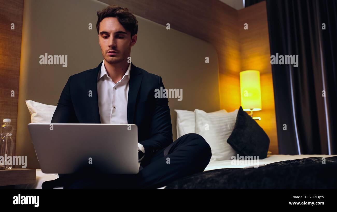 jeune homme d'affaires sérieux assis sur le lit et utilisant un ordinateur portable dans la chambre d'hôtel Banque D'Images