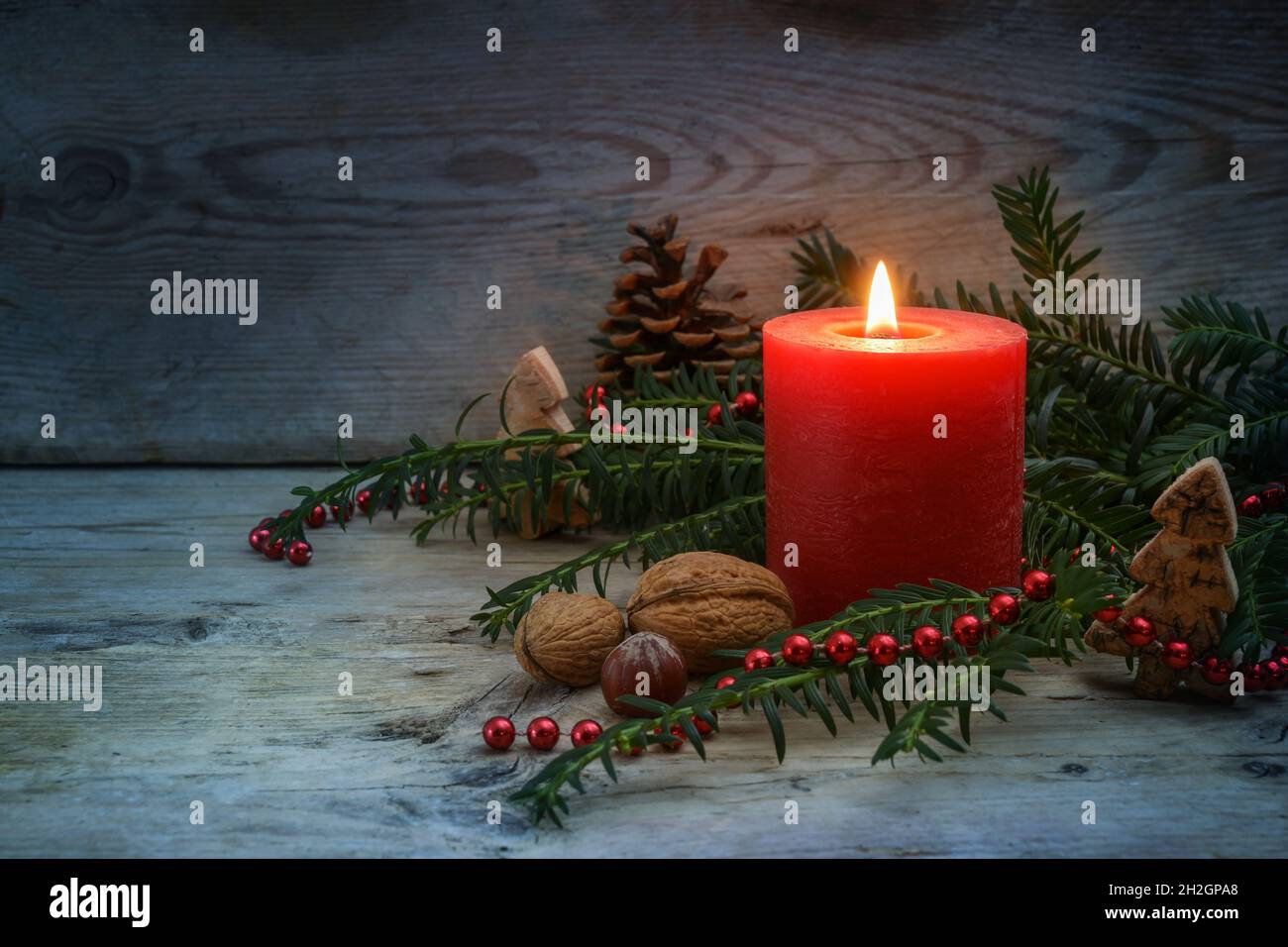 Lumière chaude sur une nuit froide d'hiver d'une bougie rouge avec une flamme, des branches de yew à feuilles persistantes, des noix et la décoration de Noël sur une planche de bois rustique gris bleu Banque D'Images