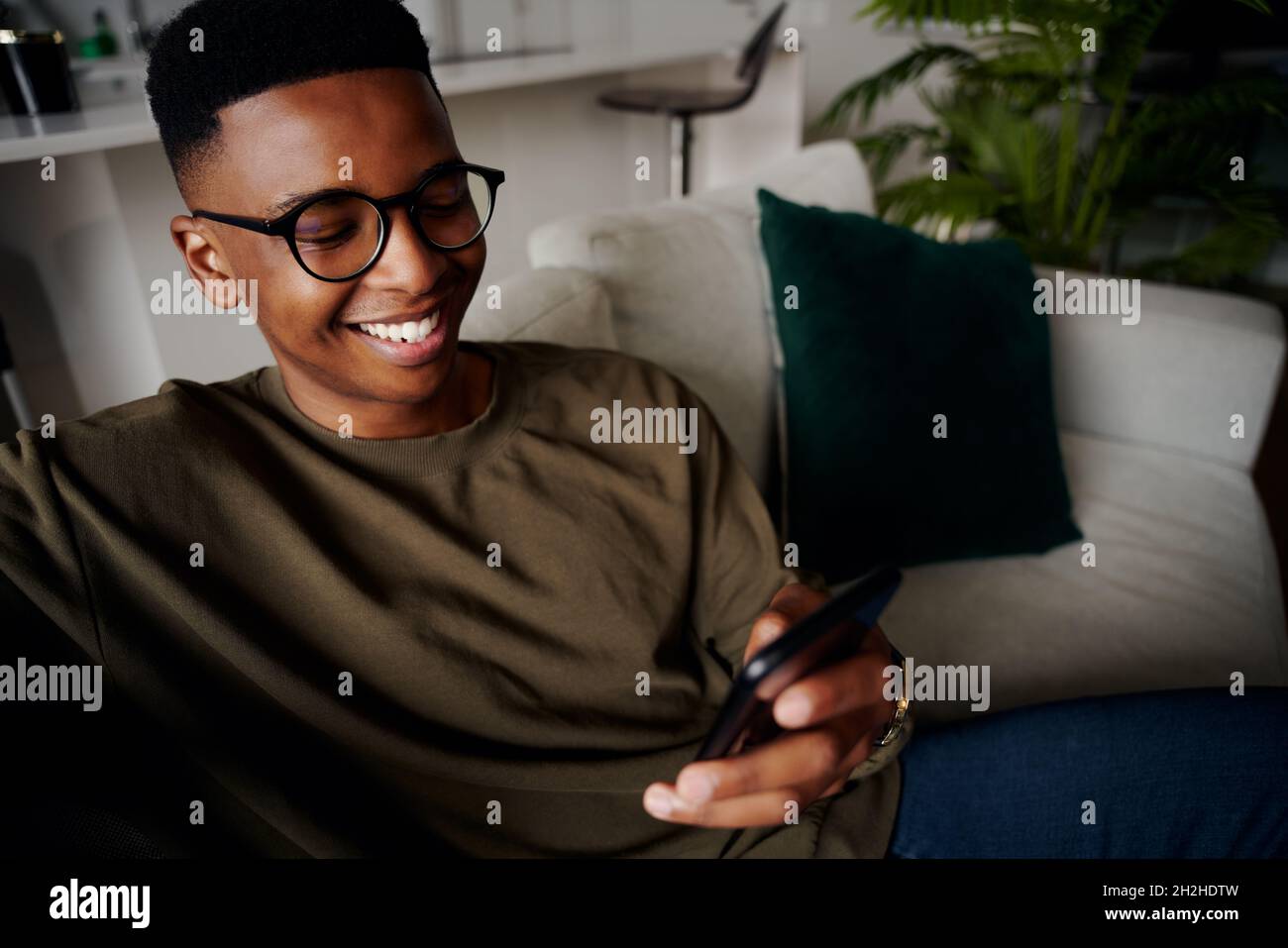 Jeune homme noir adulte décontracté, souriant tout en envoyant des SMS sur le smartphone sur le canapé Banque D'Images