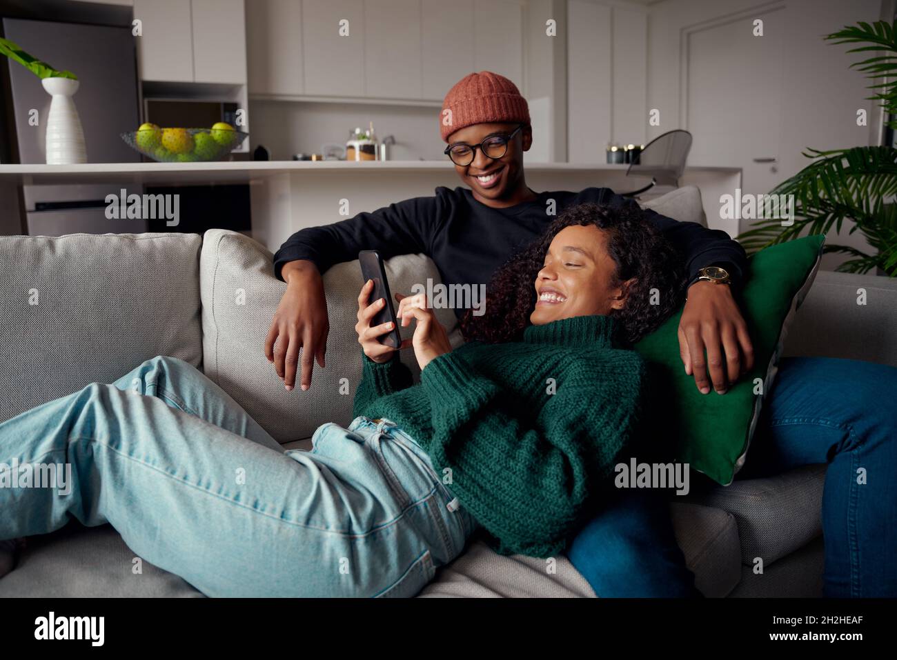 Couple multiethnique riant, regardant le texte sur le téléphone tout en étant sur le canapé dans l'appartement moderne Banque D'Images