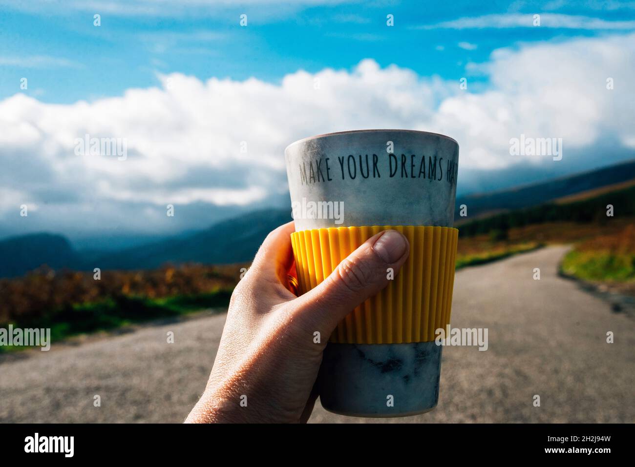 Une main tenant une tasse de voyage avec les mots font vos rêves sur elle.Route ouverte et montagnes en arrière-plan.Suivez vos rêves.Source d'inspiration Banque D'Images