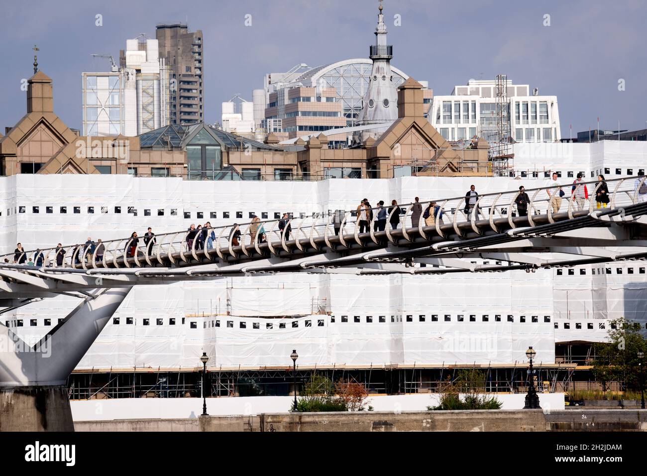 Des bâches dissimulent des travaux de construction sur des immeubles de bureaux au bord de la rivière, tandis que les Londoniens et les visiteurs de la capitale marchent sur le pont du millénaire qui traverse la Tamise, le 19 octobre 2021, à Londres, en Angleterre.La plus récente traversée de la rivière de Londres depuis plus de 100 ans a coïncidé avec le Millenium en 2000.Elle a été achevée à la hâte et ouverte au public le 10 juin 2000, quand environ 100,000 personnes l'ont traversée pour découvrir que la structure oscille tellement qu'elle a été forcée de fermer 2 jours plus tard.Au cours des 18 prochains mois, les concepteurs ont ajouté des amortisseurs pour arrêter son oscillation, mais elle est déjà fragile Banque D'Images