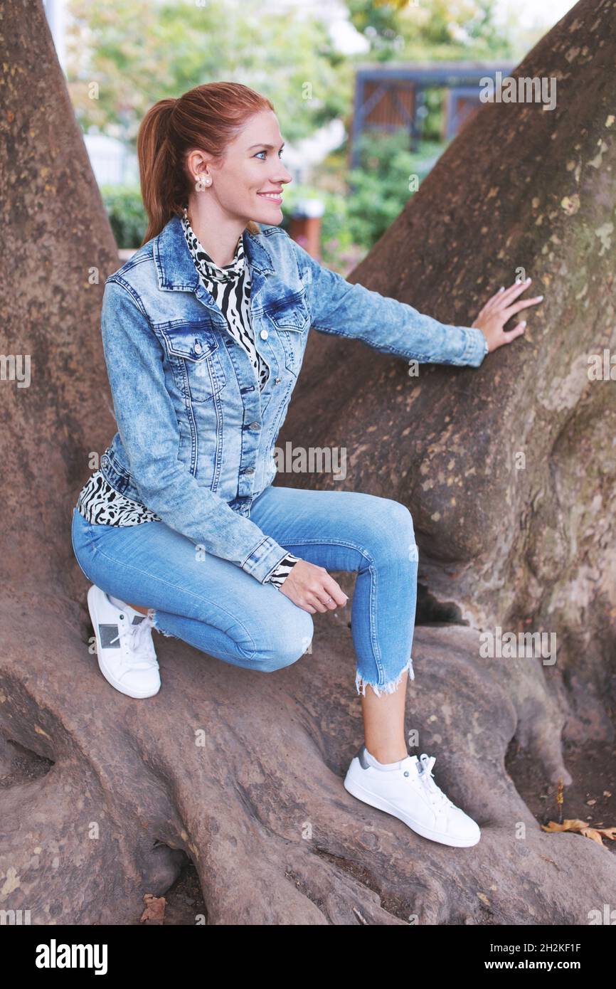 Jeune femme caucasienne à tête rouge posant sur l'arbre et regardant loin Banque D'Images