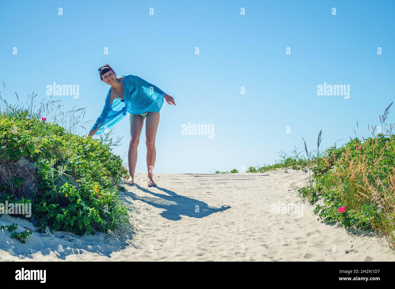 Jeune belle fille en bikini, pareo envelopper et lunettes de soleil sur la tête posant sur le sable, choisir la plante, sourire et regardant l'appareil photo, touriste femelle en vacances Banque D'Images