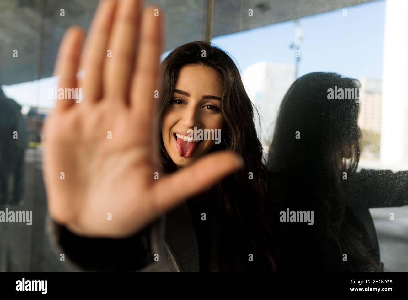 Femme qui colle la langue tout en faisant signe d'arrêt devant le mur de verre Banque D'Images