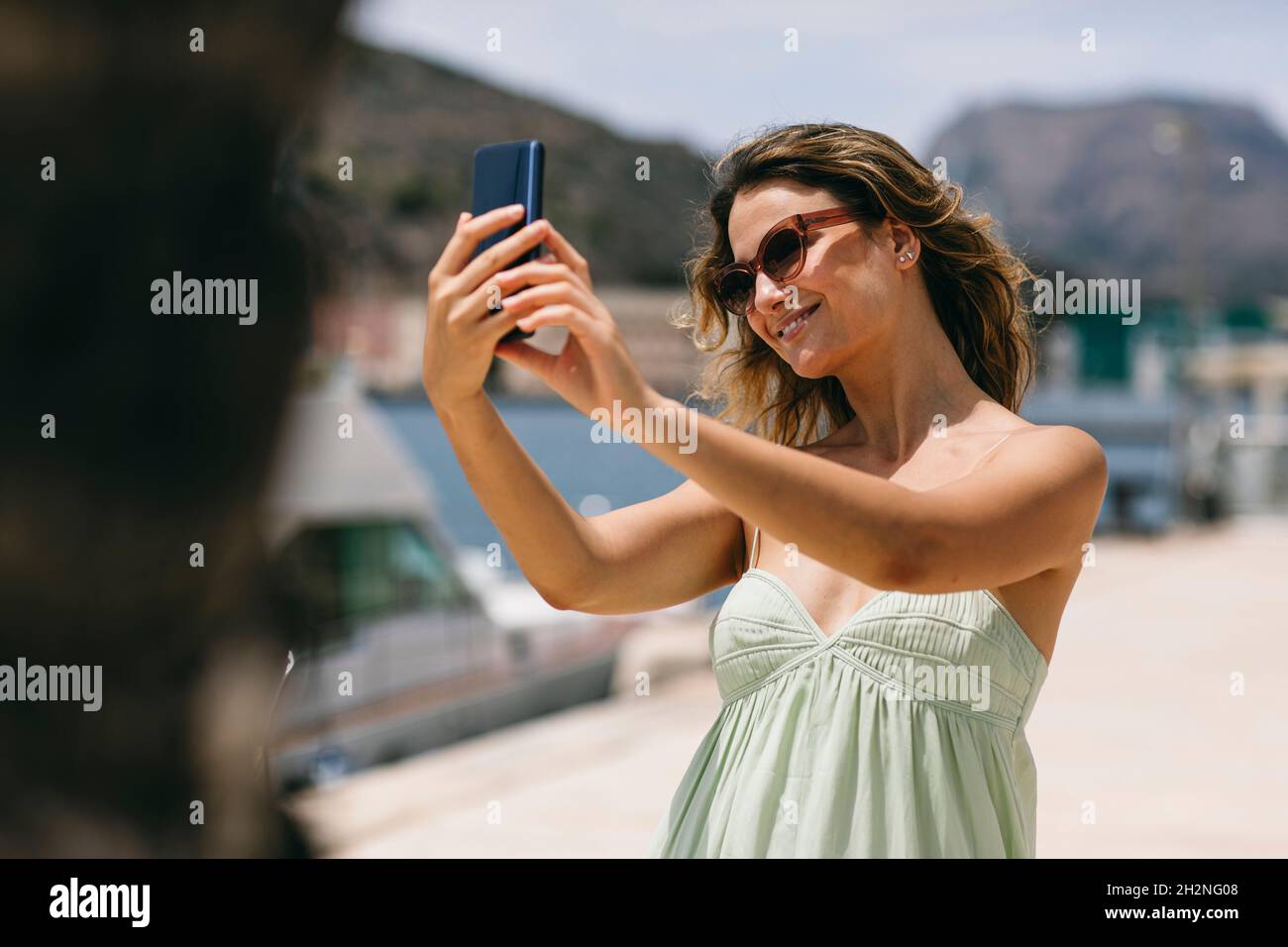 Femme souriante avec des lunettes de soleil emportant le selfie par le biais d'un smartphone Banque D'Images