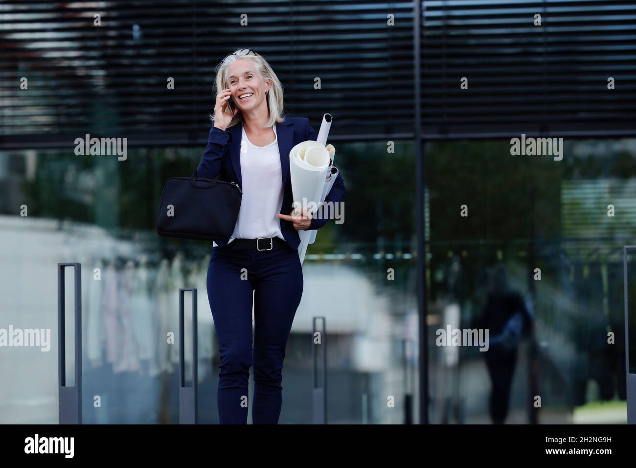 Femme d'affaires avec des plans-plans parlant sur un téléphone mobile devant le bâtiment de bureau Banque D'Images