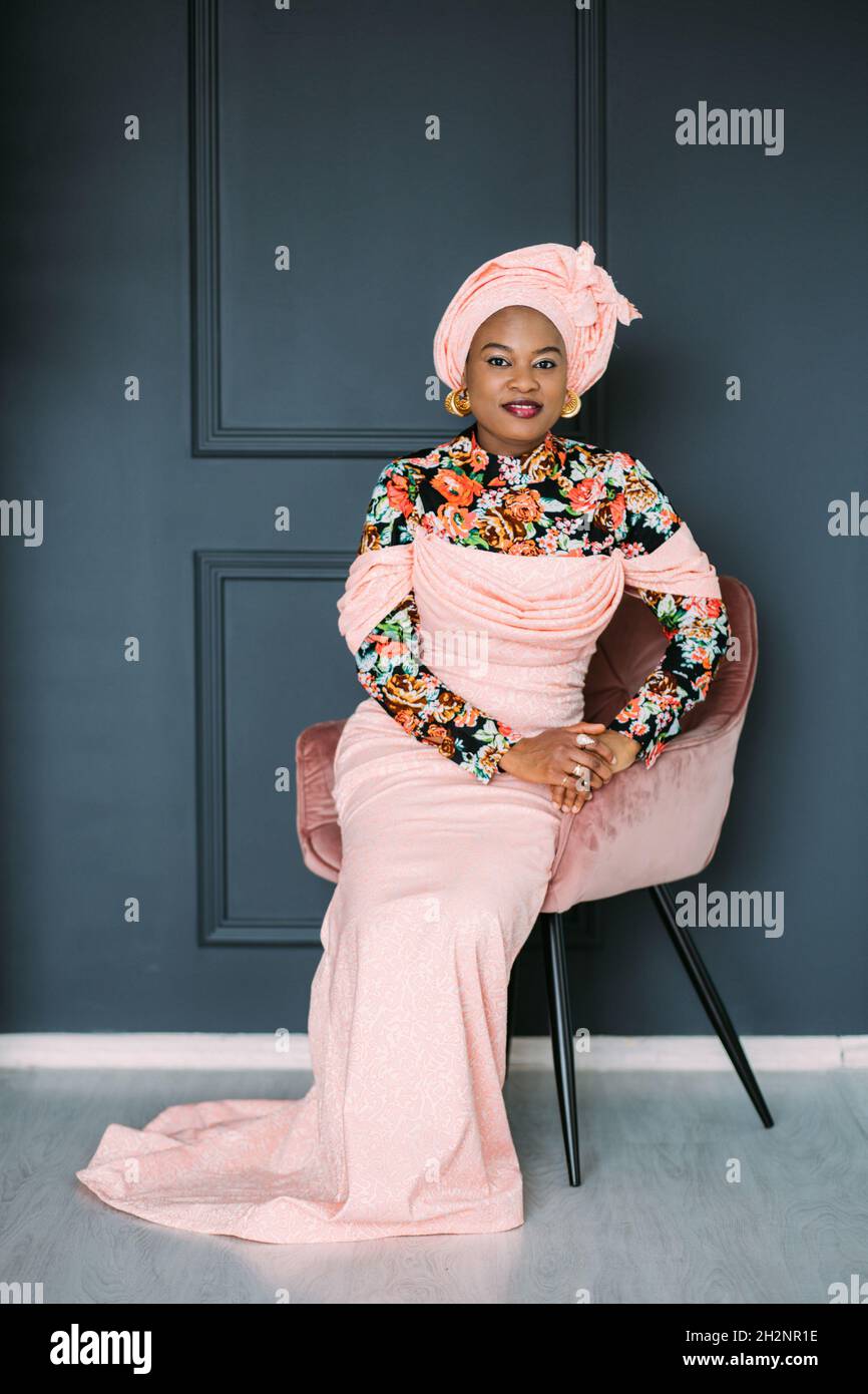 Jolie femme africaine portant une robe traditionnelle rose tendance et un  foulard, assise sur la chaise en studio et regardant l'appareil photo avec  le sourire.Fond de mur bleu foncé Photo Stock -