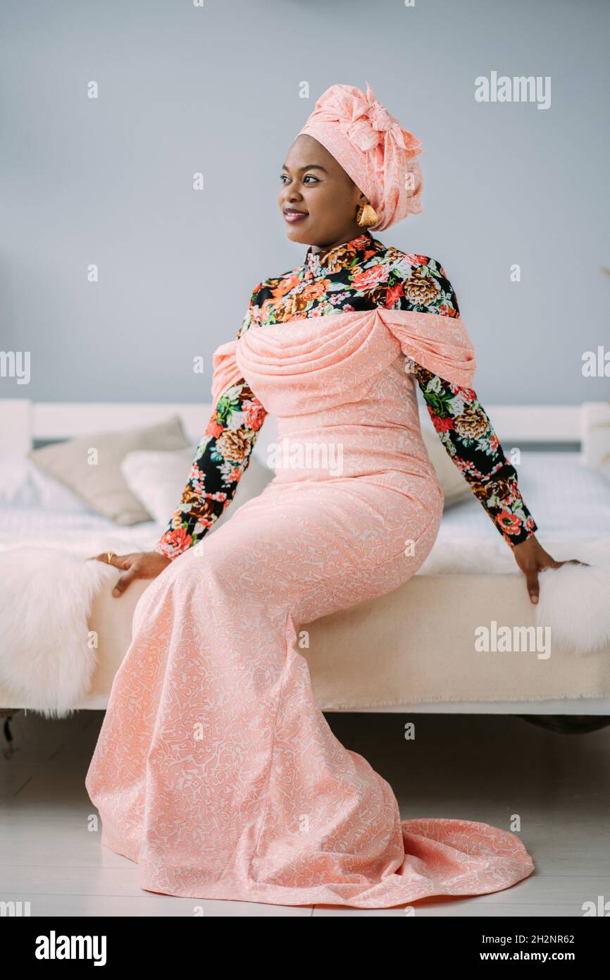 Jeune belle femme africaine portant une robe rose traditionnelle et un  foulard, boucles d'oreilles et maquillage, assis sur le lit en studio et  regardant loin Photo Stock - Alamy