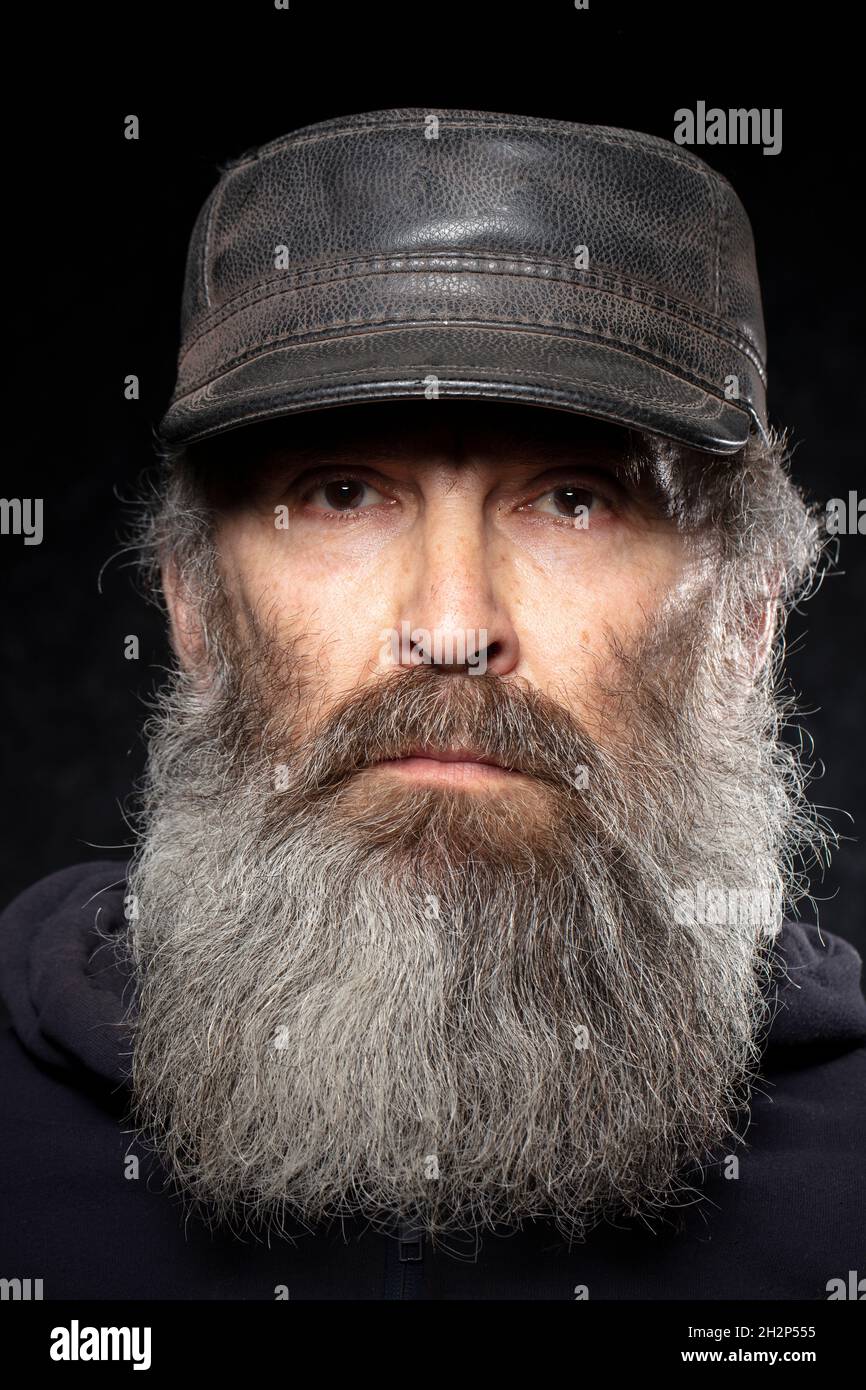 Le visage d'un homme âgé avec une barbe grise dans un chapeau en cuir sur fond noir. Banque D'Images