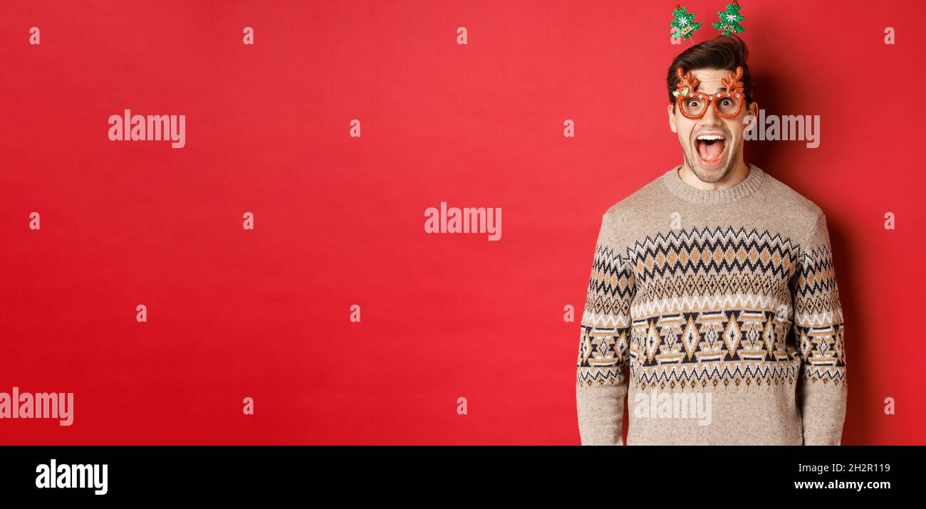 Image d'un beau homme surpris et excité, portant des lunettes de fête avec un chandail de noël, une mâchoire de chute et regardant l'offre exceptionnelle de la nouvelle année Banque D'Images