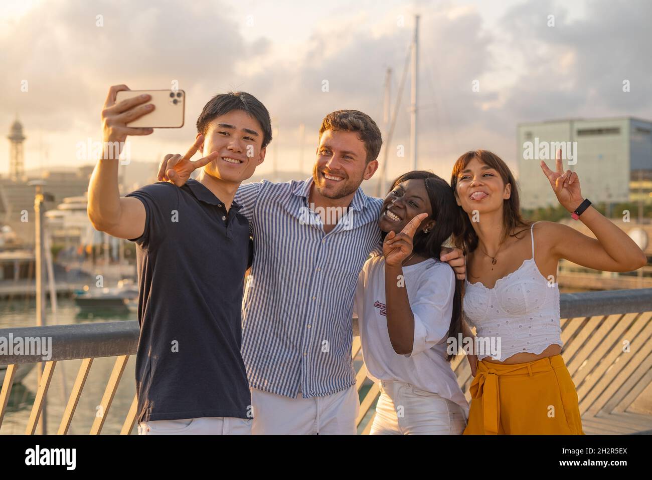 Des jeunes amis variés et insouciants dans des tenues décontractées souriant et faisant le geste de paix tout en emportant le selfie sur un smartphone pendant le temps libre ensemble sur les remblais en soirée d'été Banque D'Images