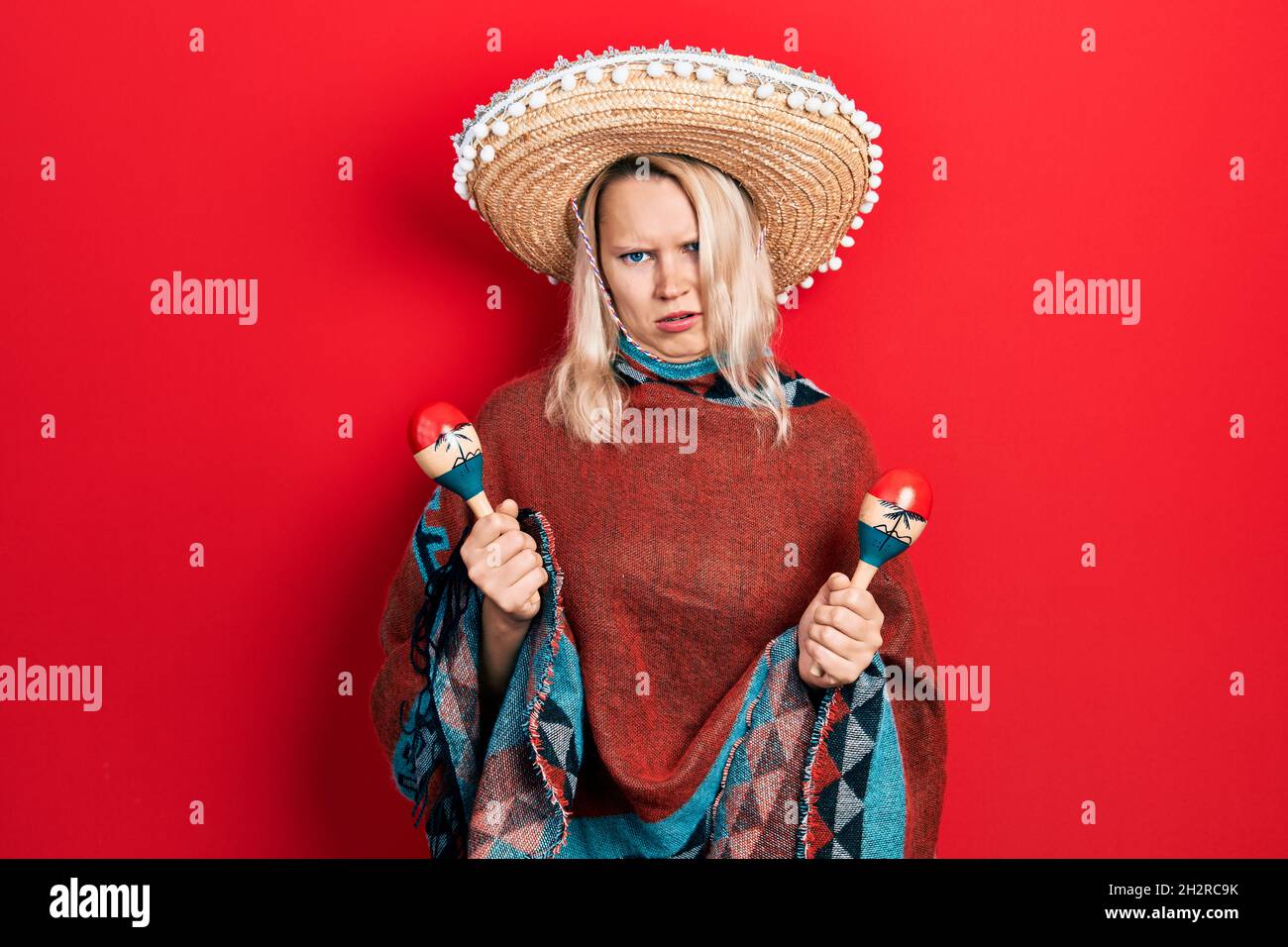 Belle femme blonde caucasienne portant poncho mexicain festif et maracas expression sans clus et confuse. Doute concept. Banque D'Images