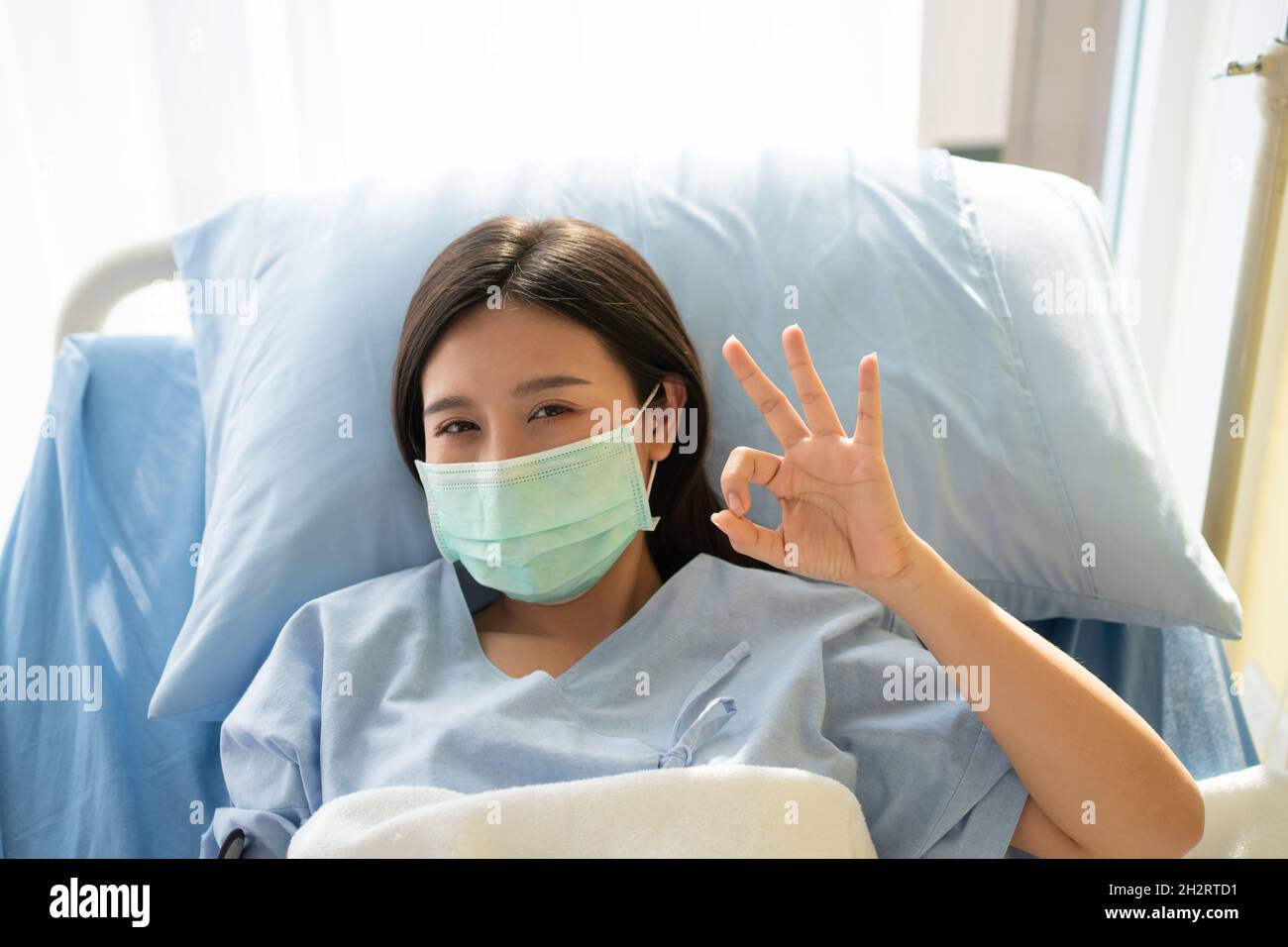 Une patiente asiatique heureuse porte un masque, repose sur le lit et affiche un symbole ok pour montrer la confiance dans le traitement.Concept de croire en traitement et Banque D'Images