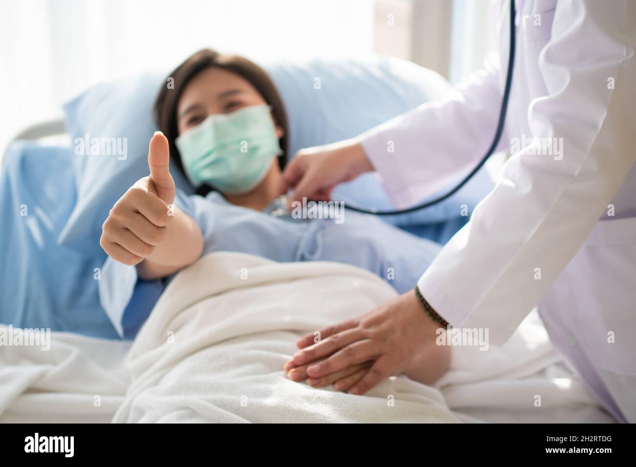 Une patiente asiatique heureuse porte un masque, se repose sur le lit et lève le pouce.Lorsqu'un médecin utilise un stéthoscope pour écouter les poumons.Concept de Banque D'Images