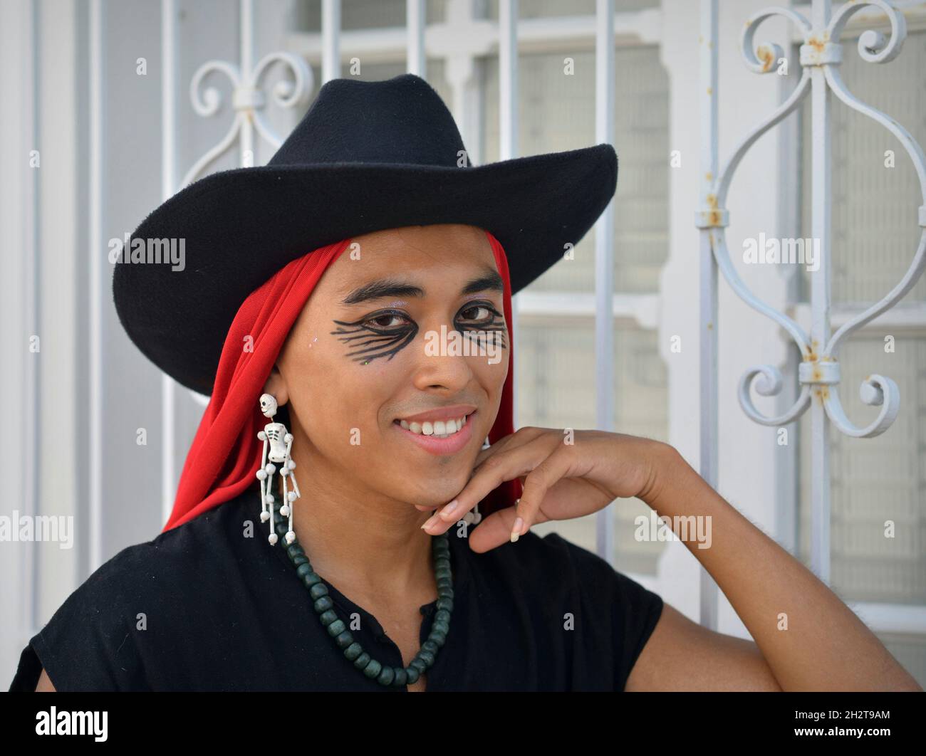 Joli jeune Latino non binaire avec chapeau noir, rouge mexicain rebozo et le squelette Day of the Dead sourit boucles d'oreilles devant la grille blanche de fenêtre. Banque D'Images