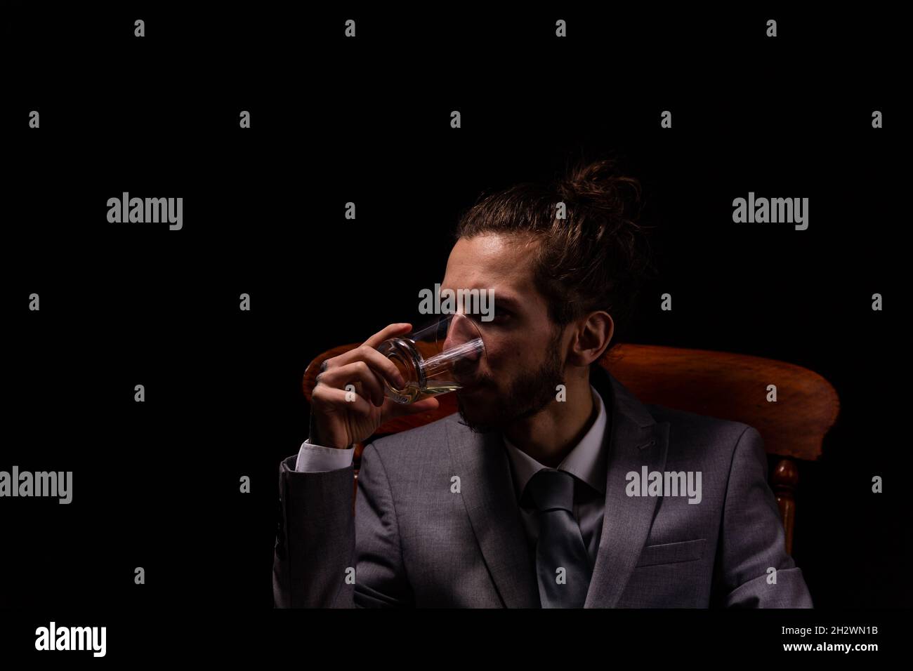 Jeune homme d'affaires en costume gris élégant, assis dans un fauteuil en bois avec éclairage d'ambiance et de mauvaise humeur, il boit une seule photo de whisky Banque D'Images