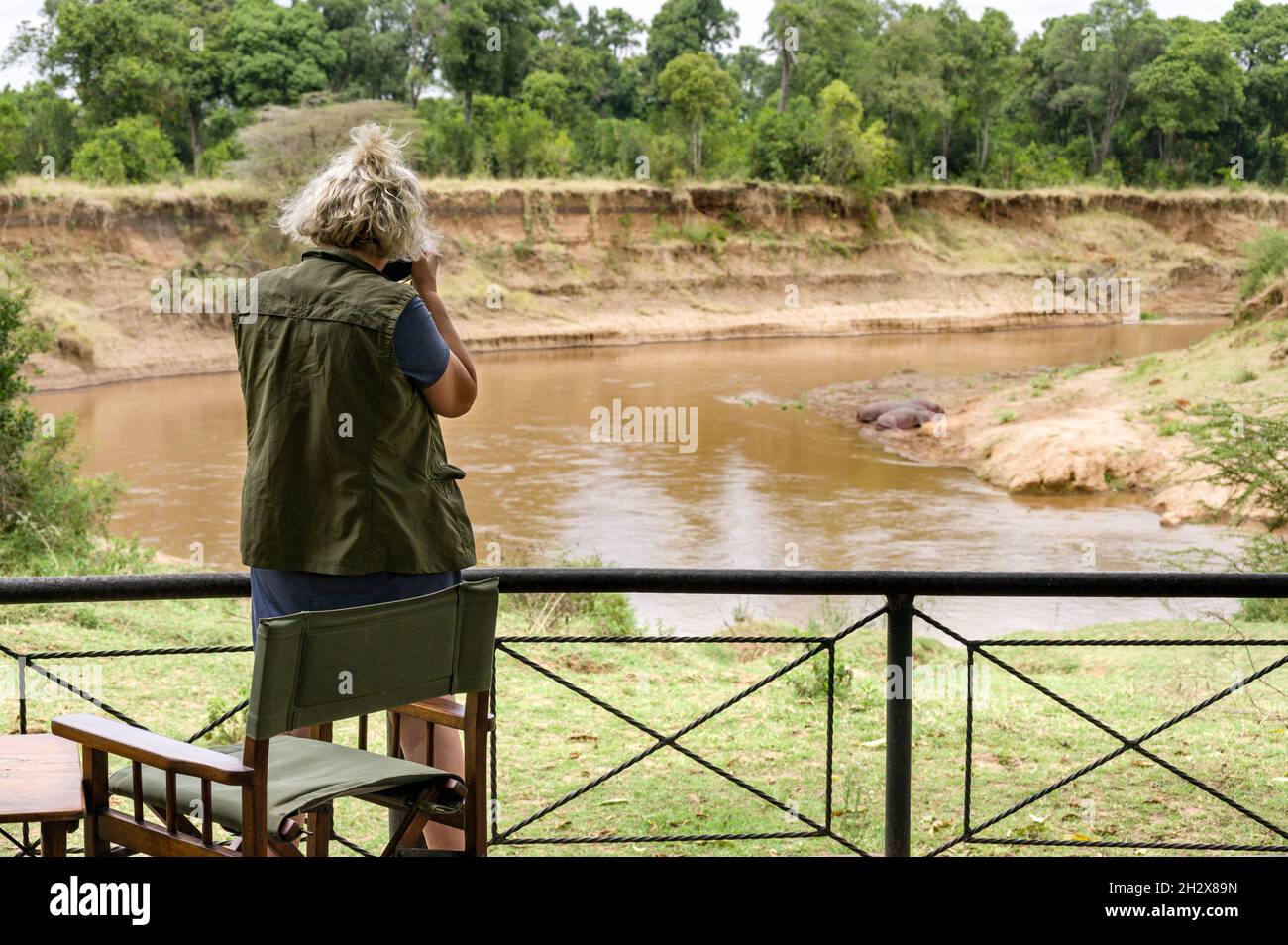Une femme caucasienne se tient près d'un garde-corps prenant des photos d'hippopotames par la rivière Mara à distance, Masai Mara, Kenya Banque D'Images