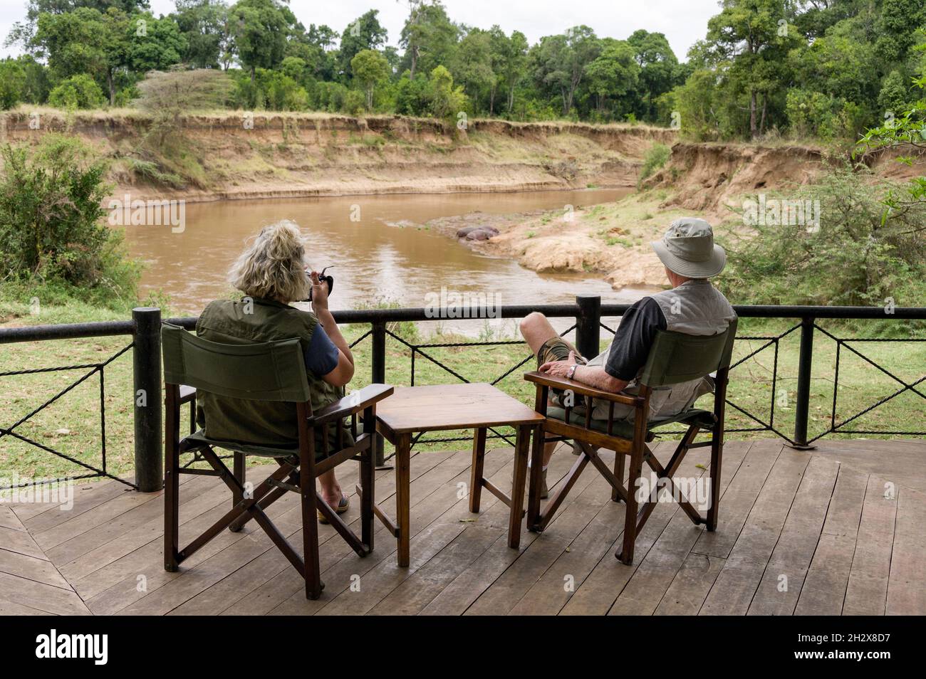 Couple caucasien assis sur la zone d'observation au camp de Governors observant les hippopotames près de la rivière Mara à distance, Masai Mara, Kenya Banque D'Images
