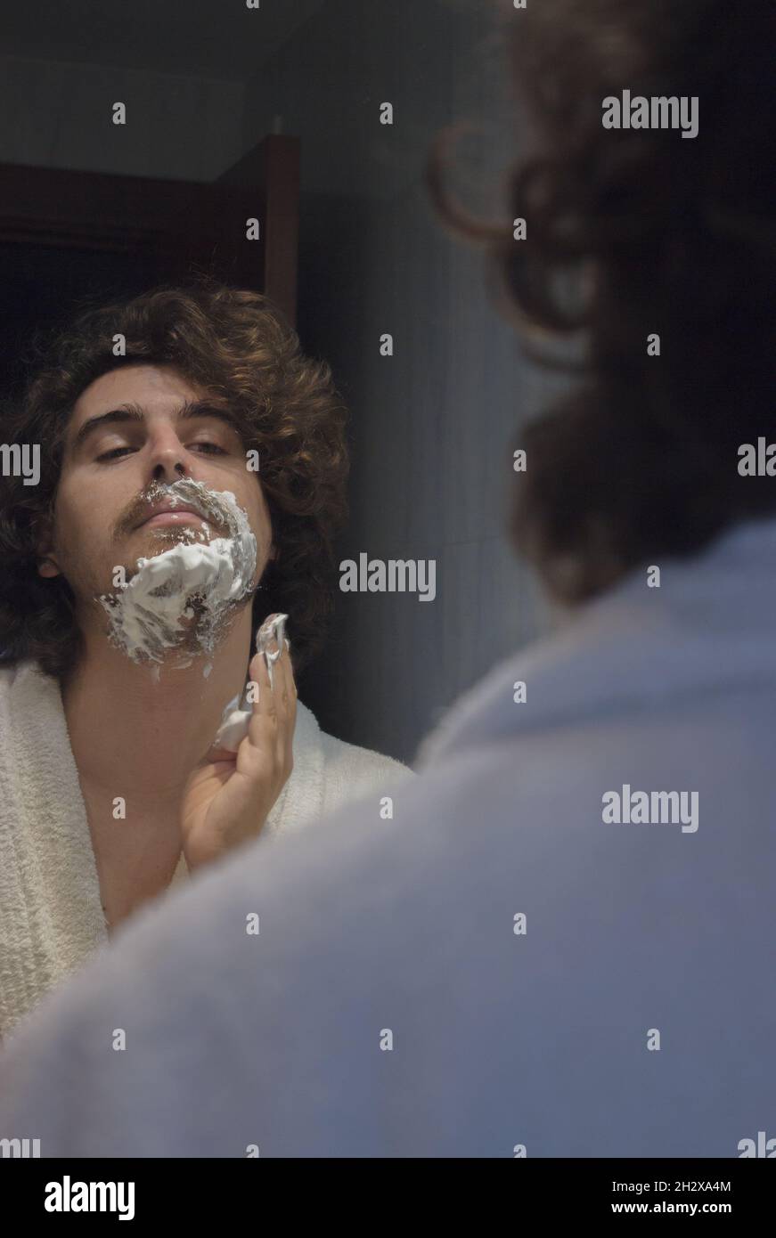 Un jeune homme en peignoir applique de la mousse de rasage sur le visage dans la salle de bains de l'appartement.Concept de spa Beauty Home. Banque D'Images