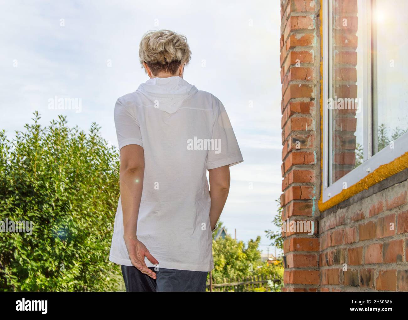 Une jeune blonde élancée avec une coupe courte tendance dans un t-shirt blanc avec capuche marche à l'extérieur, le long d'une maison avec un mur de briques rouges.Vue arrière Banque D'Images