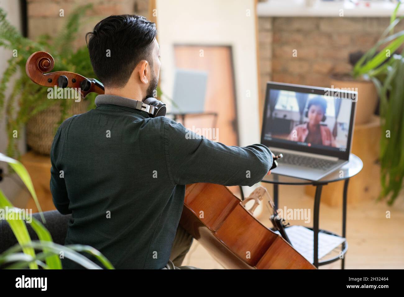 Vue arrière d'un jeune professeur de musique jouant du violoncelle devant un ordinateur portable avec un joli petit écolier à l'écran Banque D'Images