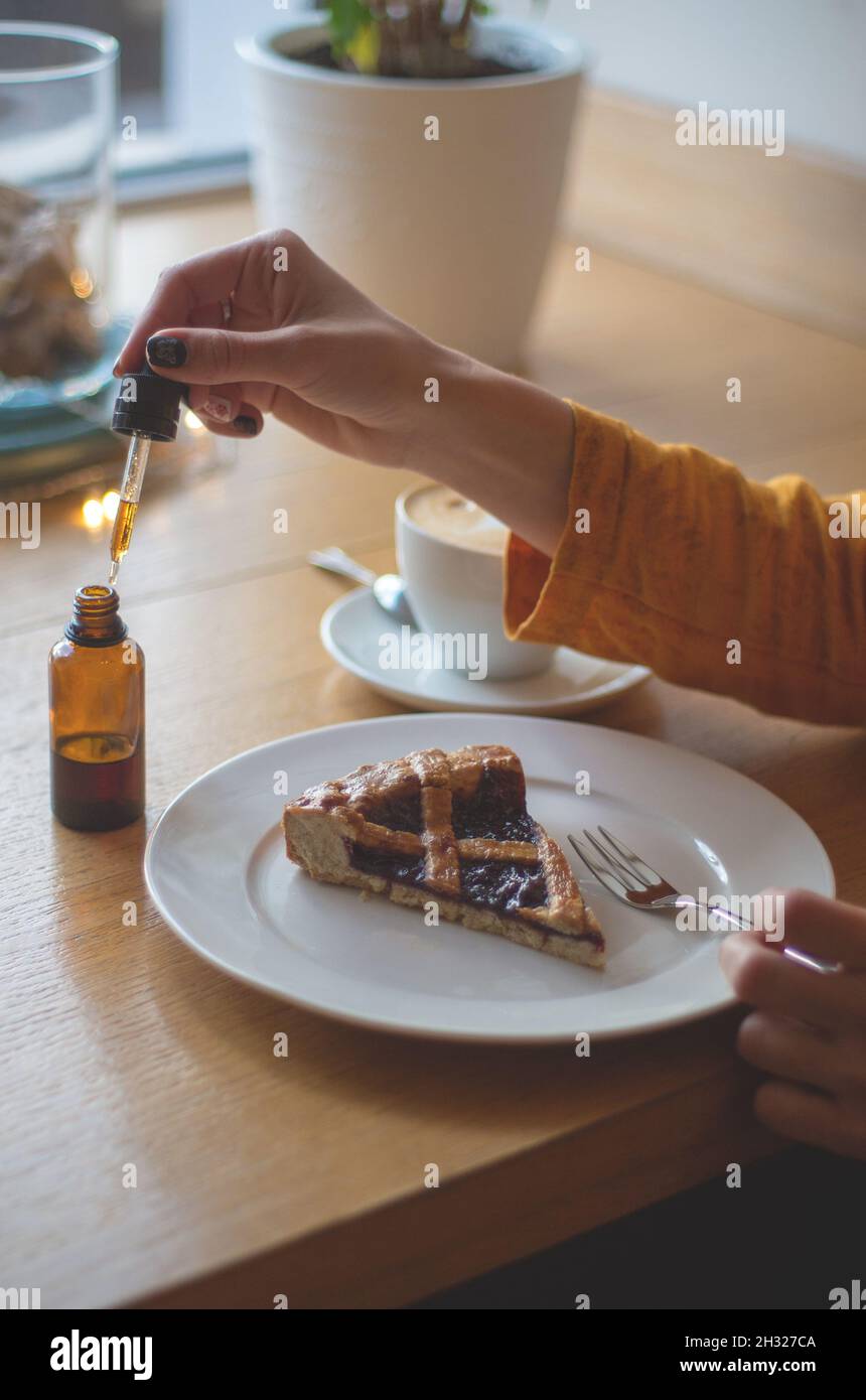Une fille dans une veste jaune prend le petit déjeuner dans un café.Les mains des femmes ajoutent du THC et de l'huile de CBD à la tarte aux cerises.Le concept de l'utilisation de la marijuana médicale. Banque D'Images