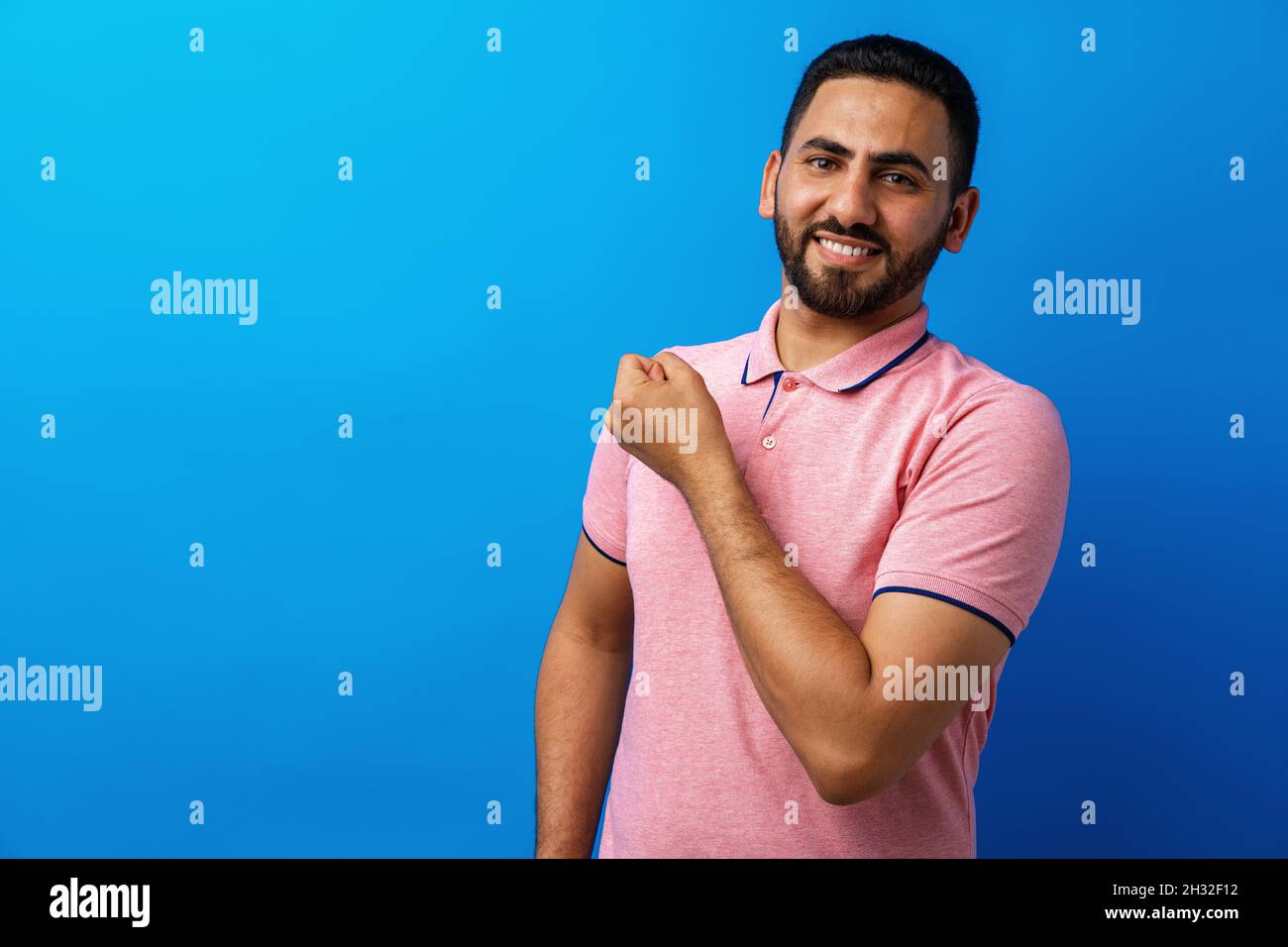 Homme hispanique avec barbe portant un t-shirt décontracté montrant les muscles des bras sur fond bleu Banque D'Images