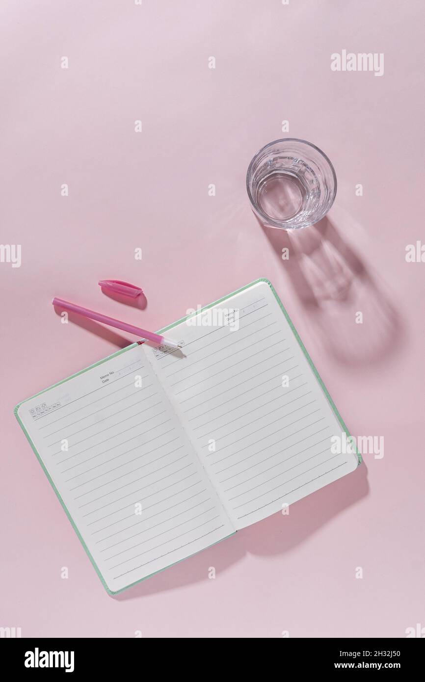 Rose espace de travail féminin encore vie avec journal ouvert ou carnet, stylo rose et un verre d'eau douce saine dans une vue de dessus en bas plan avec Banque D'Images