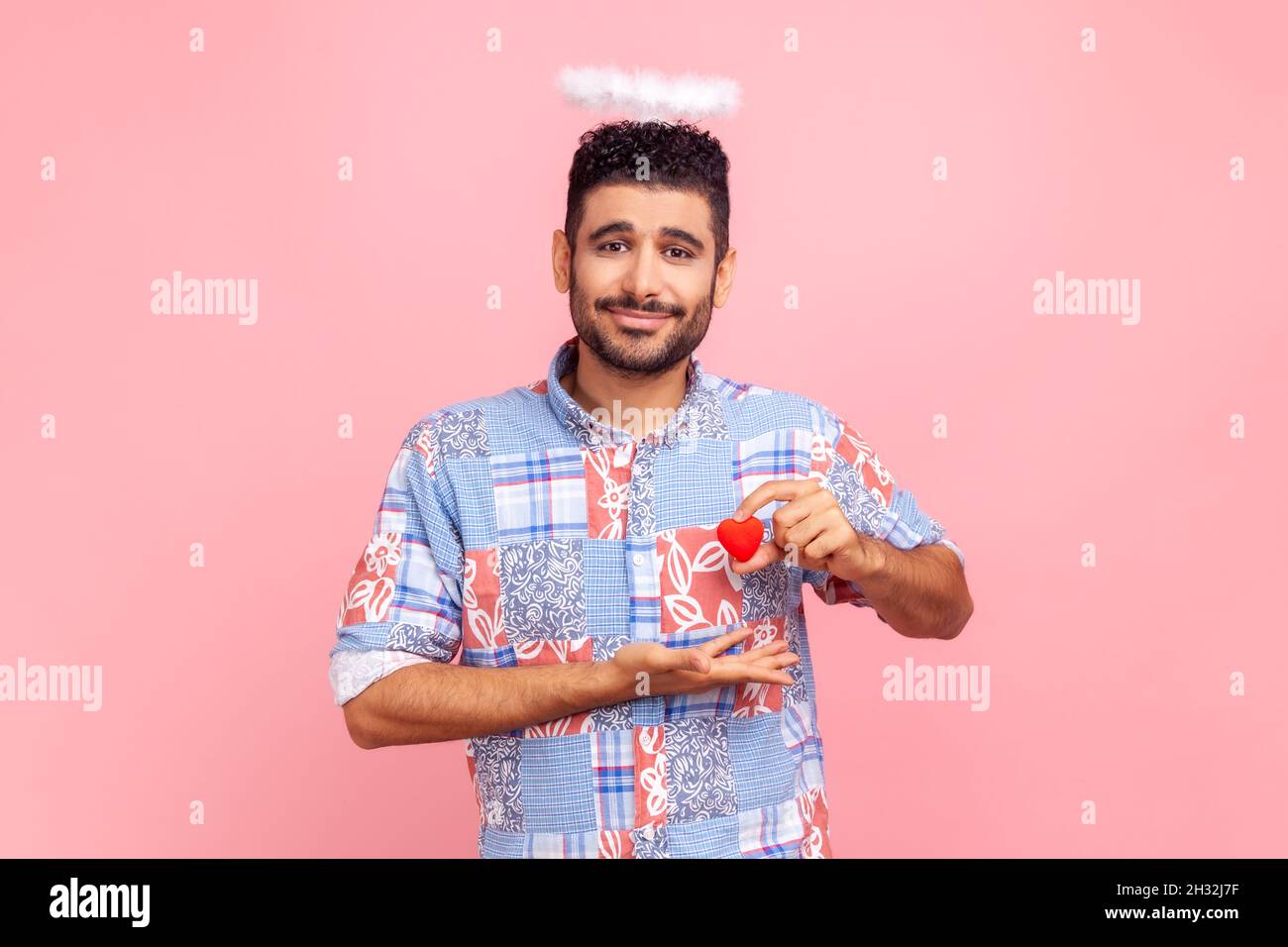 Portrait d'un bel homme barbu avec nimb au-dessus de la tête tenant un petit coeur rouge dans les mains, regardant l'appareil photo avec amour, portant une chemise bleue décontractée.Studio d'intérieur isolé sur fond rose. Banque D'Images